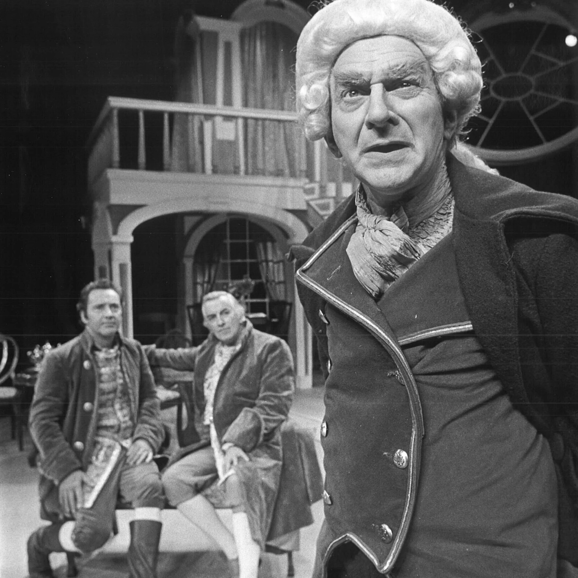 A man wearing a long coat and a white wig from the 1800's is staring off into the distance while two men are sitting on a bench behind him looking at him.