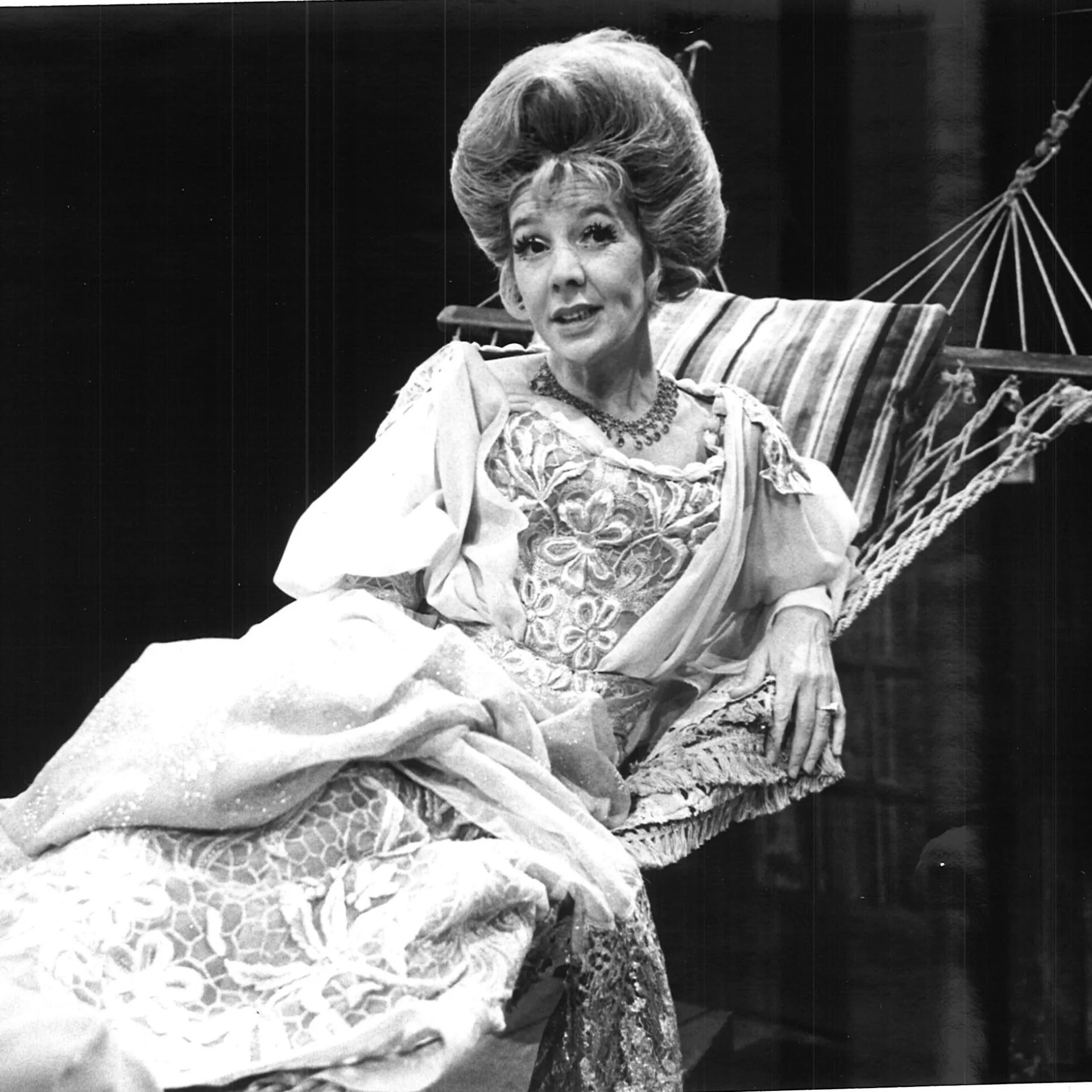 A woman dressed as a 1950's housewife, is lounging on a hammock looking ahead.