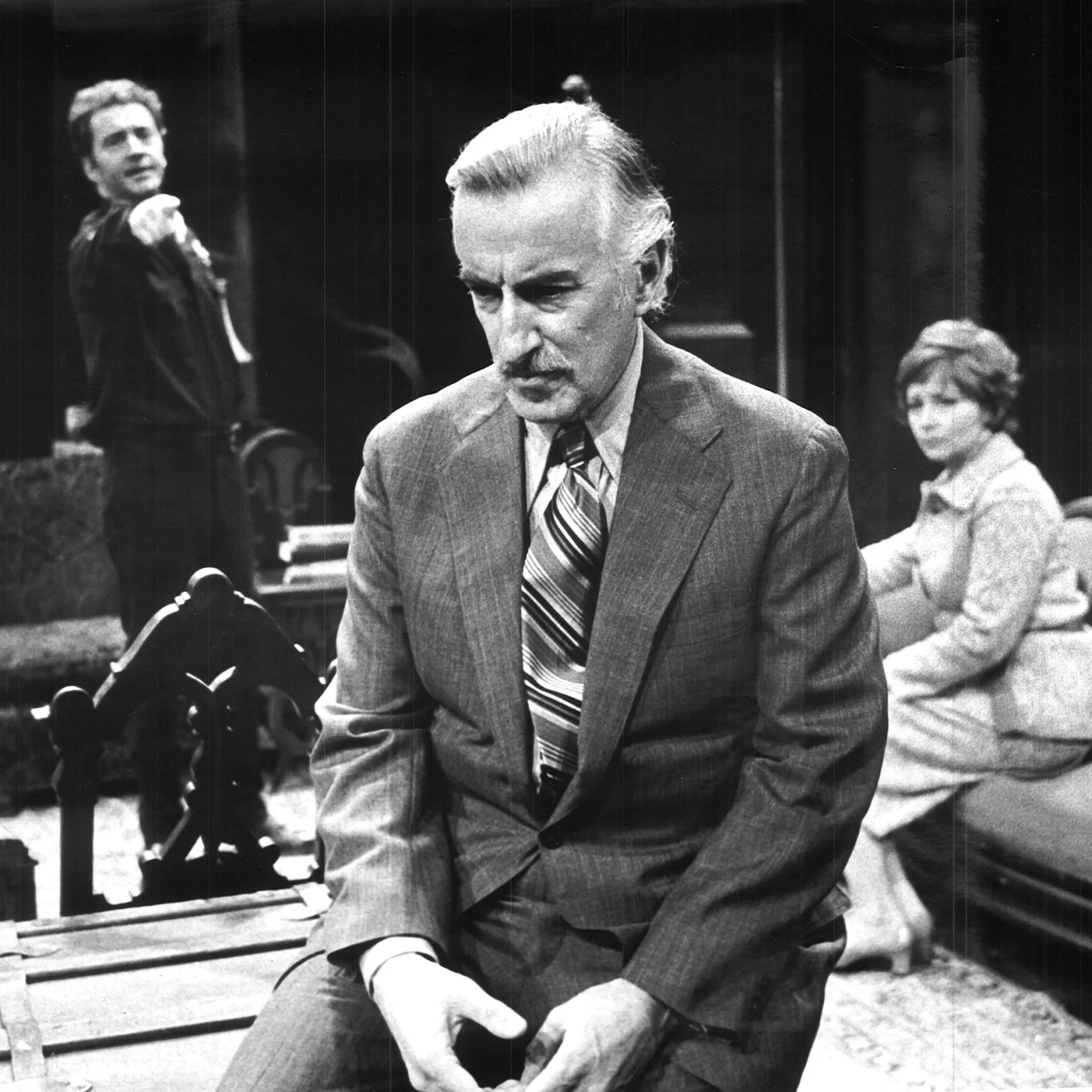 A man is seen sitting and looking down while another man in the background is standing up and pointing at him, across from him a woman is sitting down and watching him.