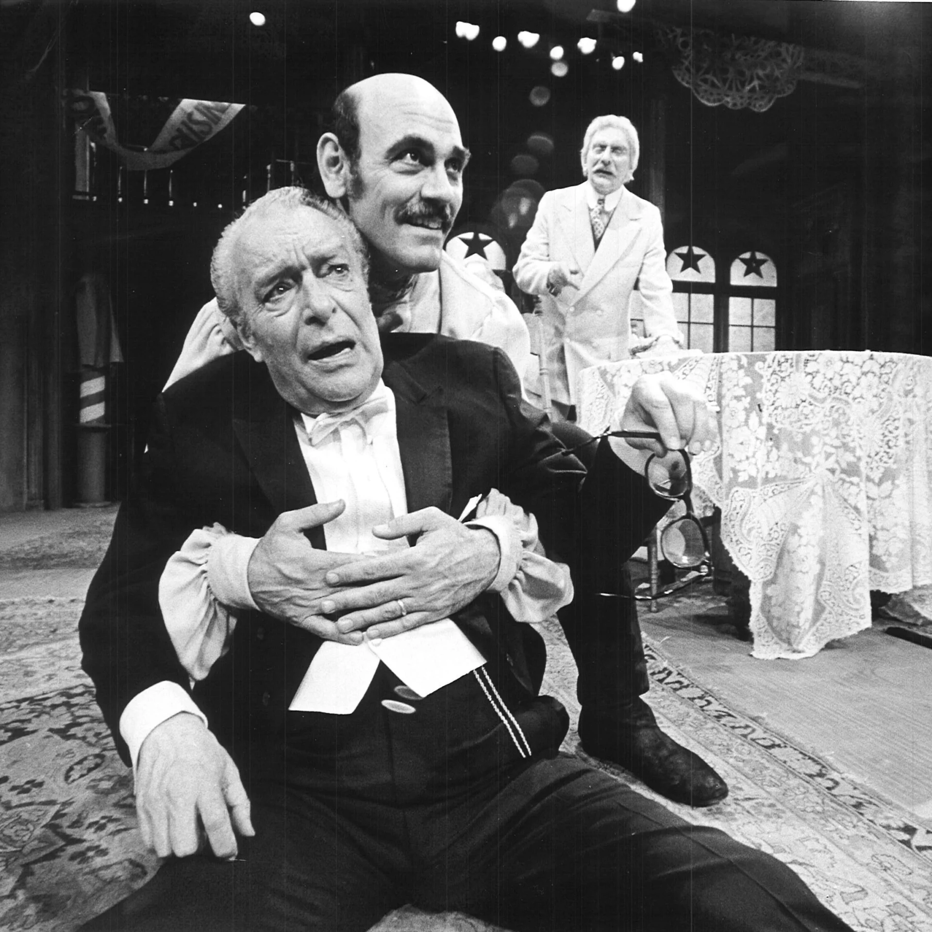 A Man is giving another man the Heimlich maneuver while another man is shouting and pointing in the background.