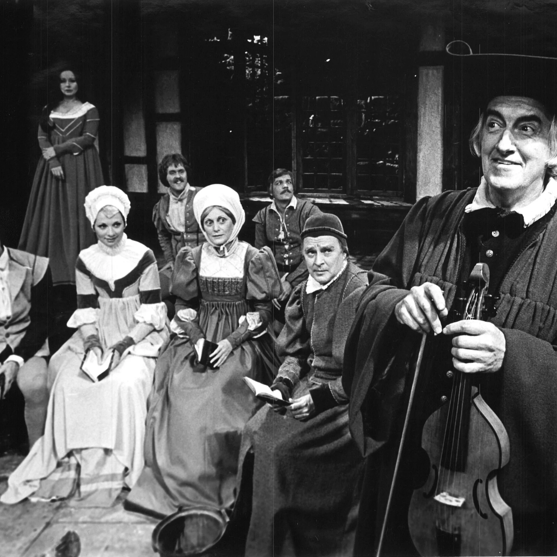 A group of woman wearing dresses from the 1700's are sitting down and watching a man who's holding a violin