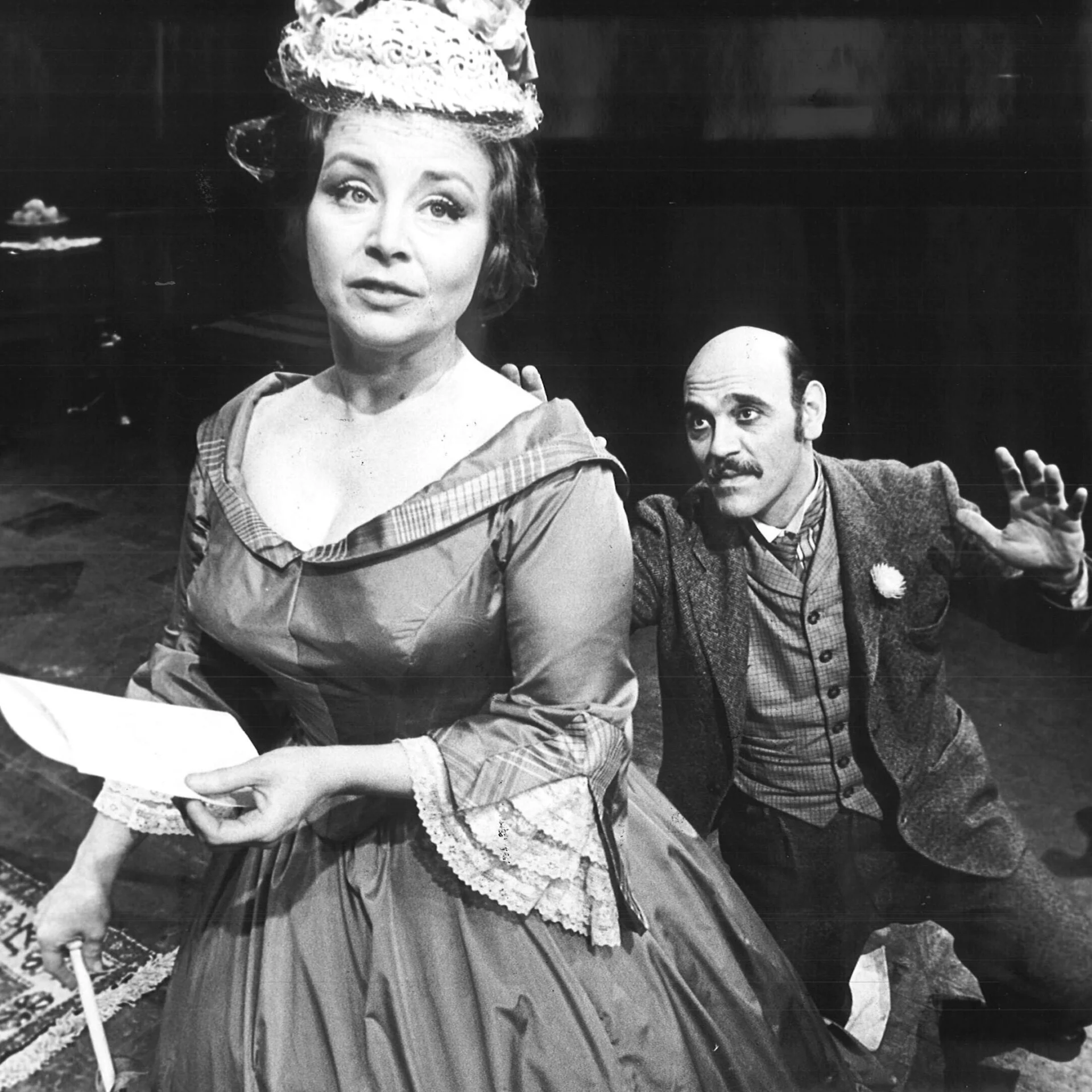 A woman dressed in a Victorian styled dress is looking away while a man is begging on his knees behind her.