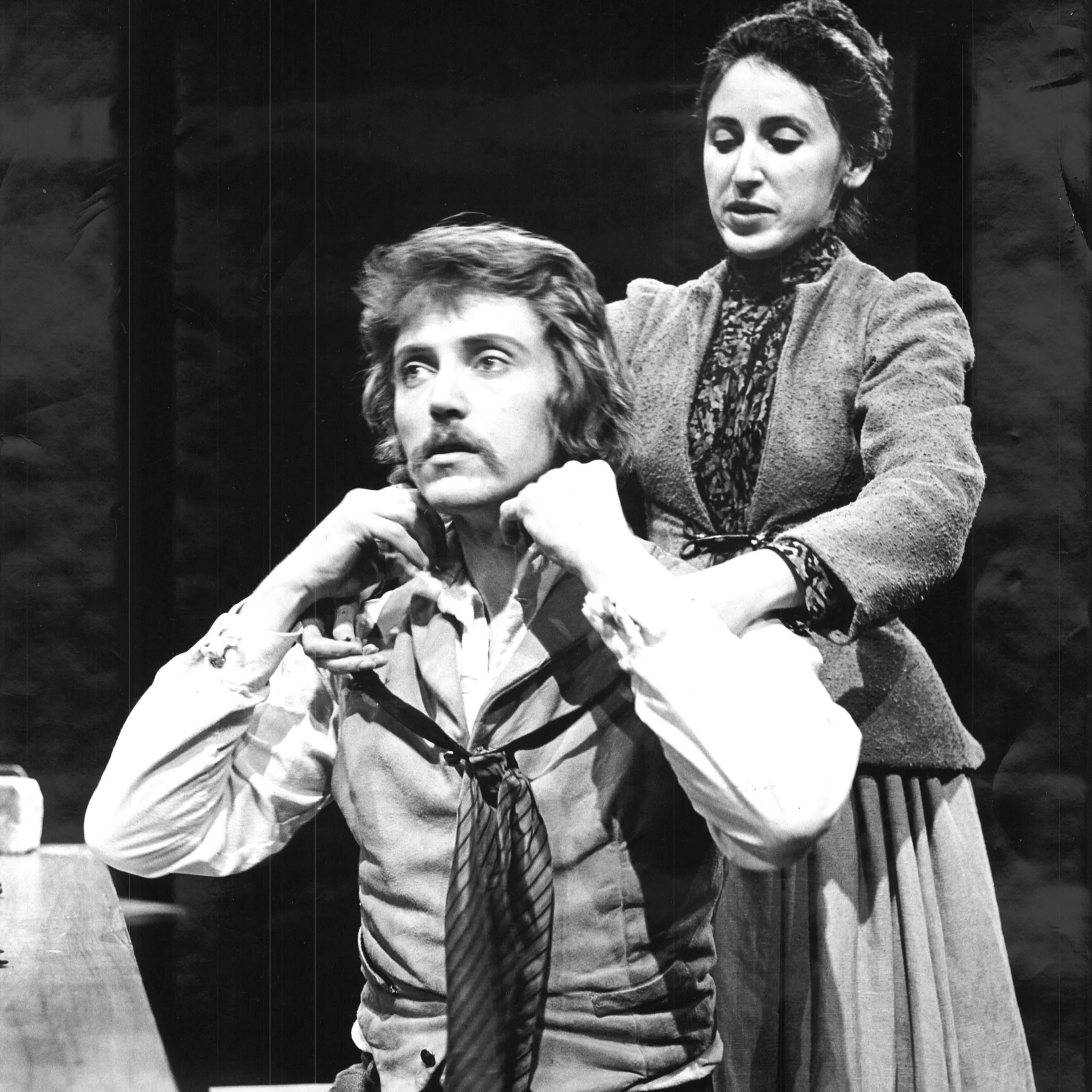 A man is sitting down in a chair while a woman is standing behind him and tying his tie.