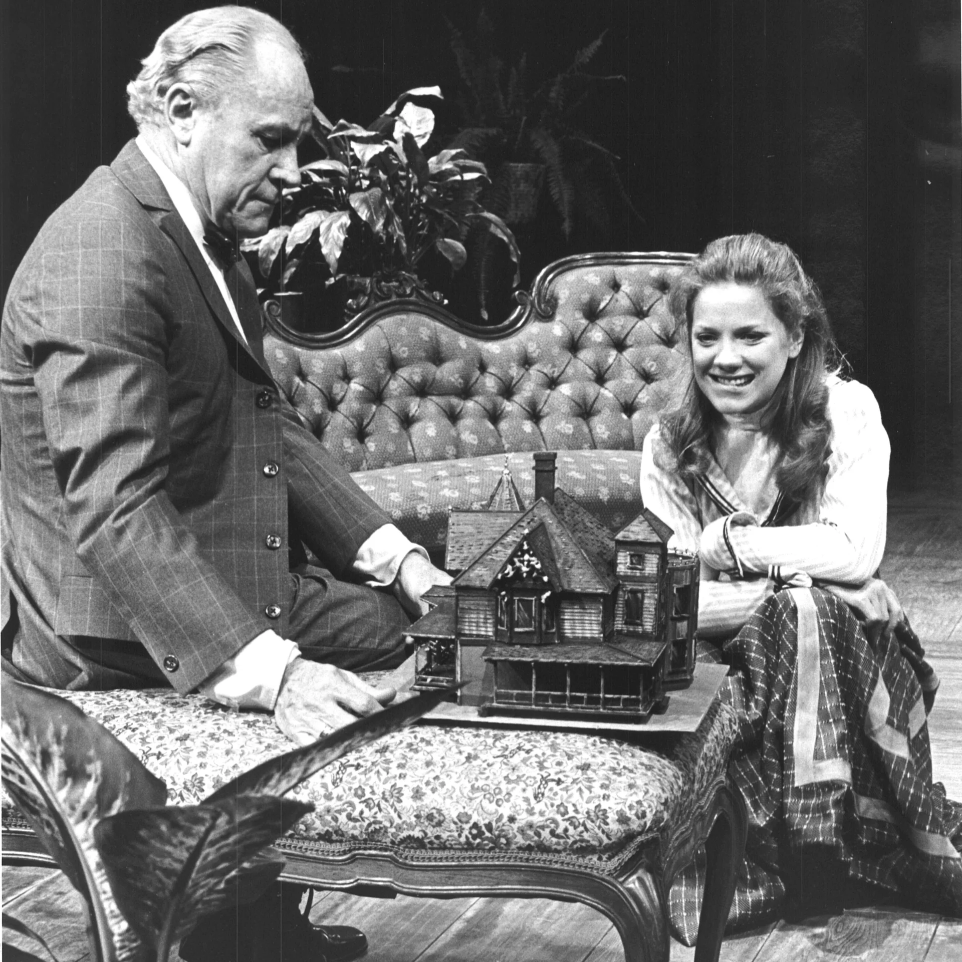 An older man and a younger woman are seen crouching over a tiny house replica and the woman is smiling excitingly.