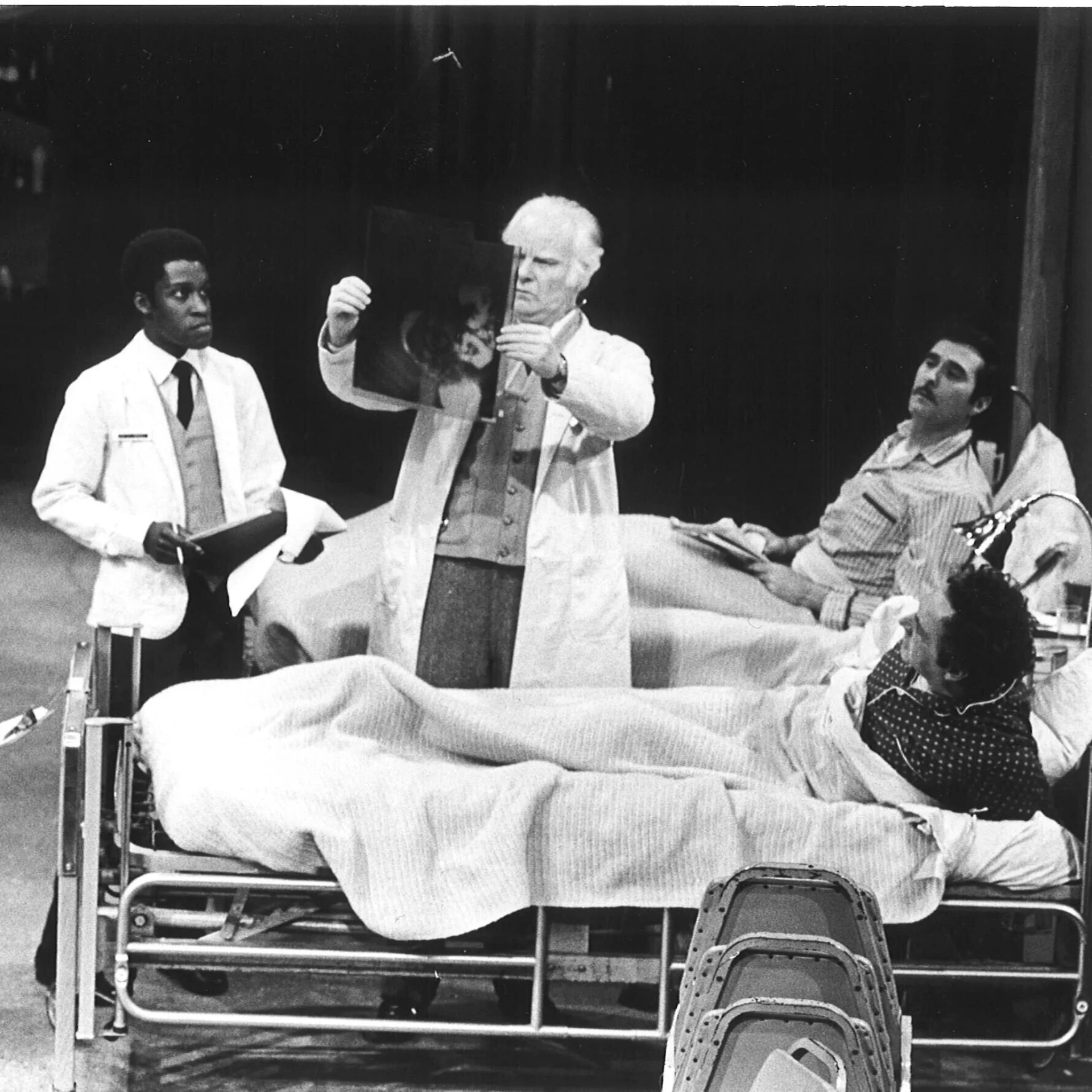 Two men are sitting in hospital beds with two doctors standing in between them and assisting them.