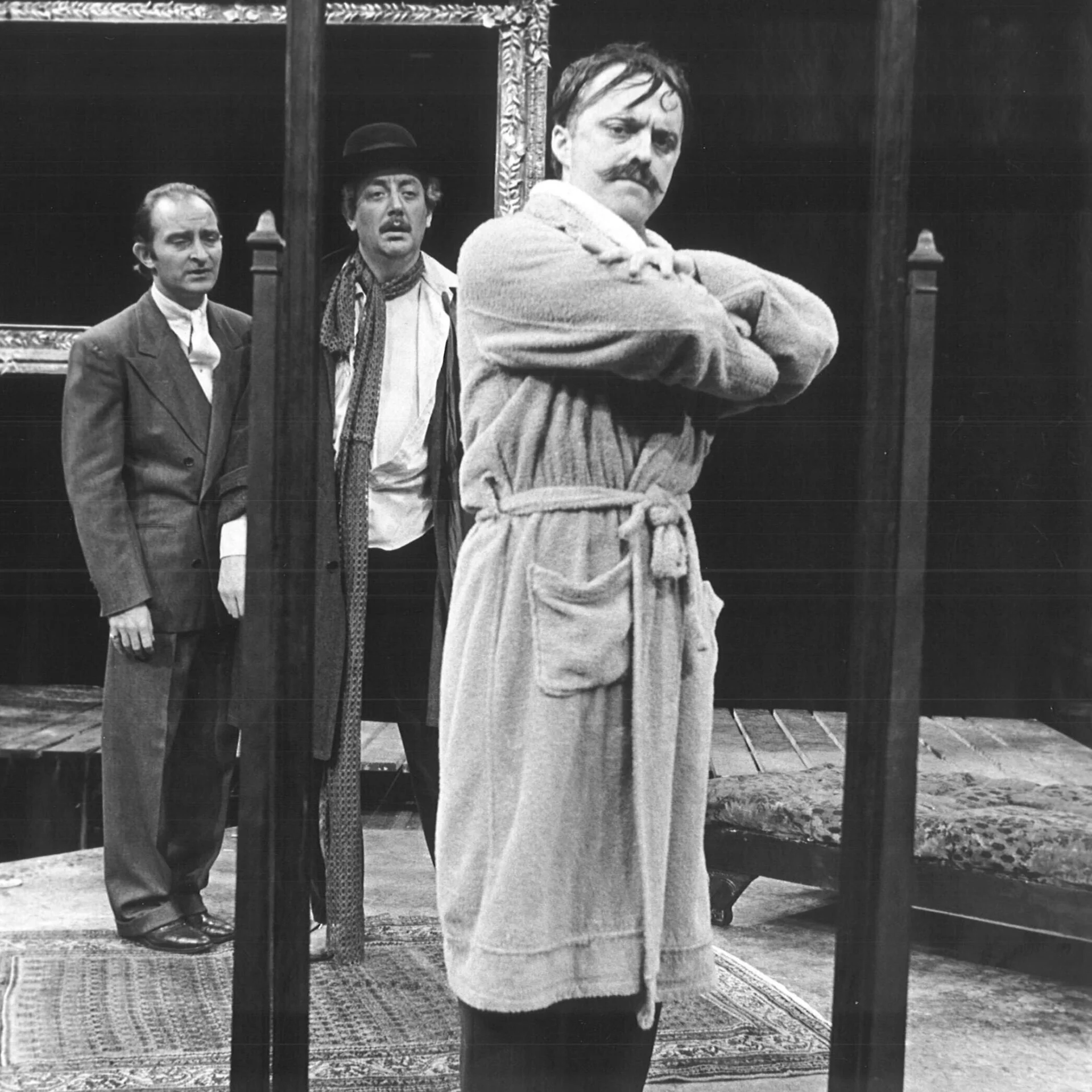 A man is standing in a bathrobe with his arms crossed while two other men are watching him in the background