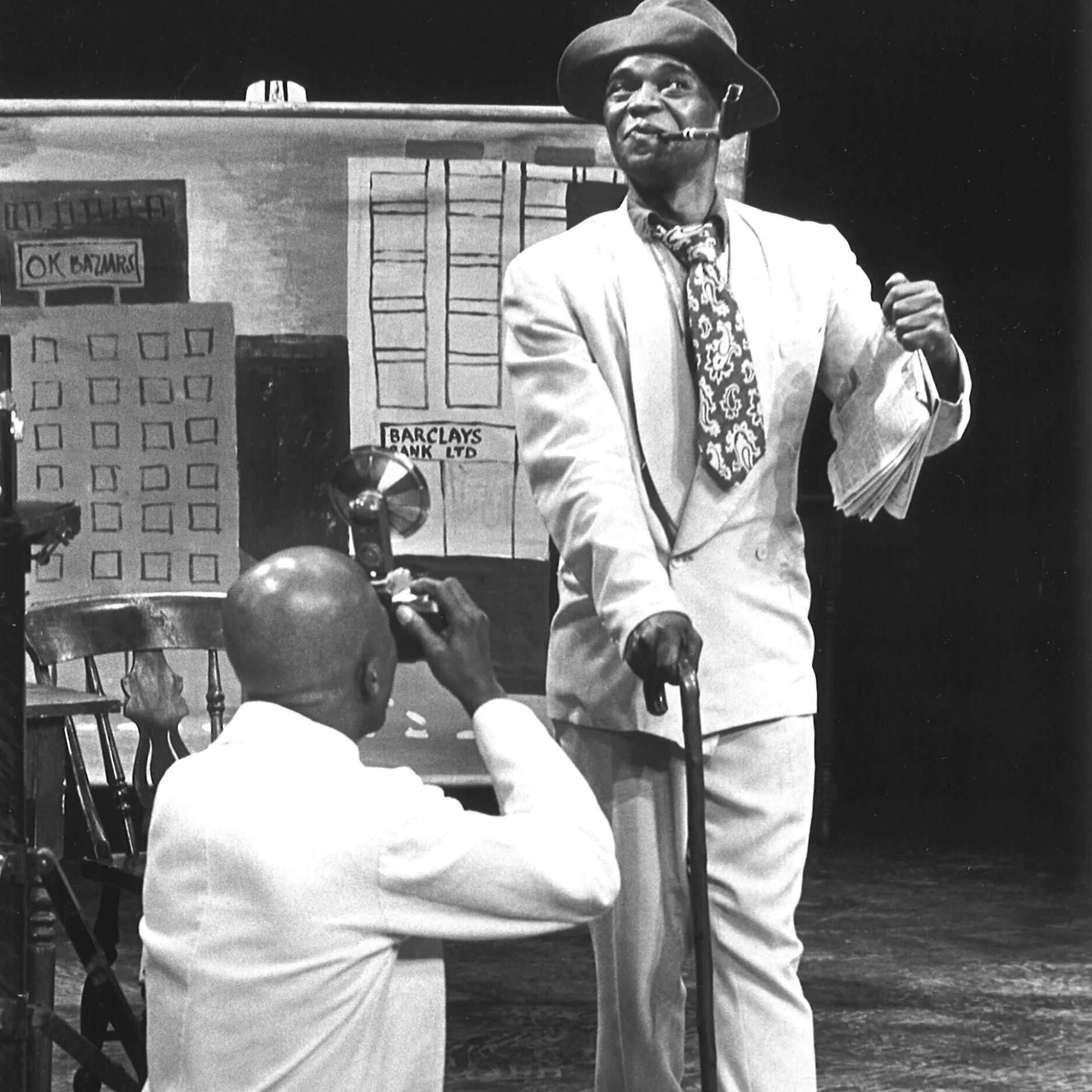 One man is standing holding a cane while he poses for a picture and another man is crouched down and taking a picture of him