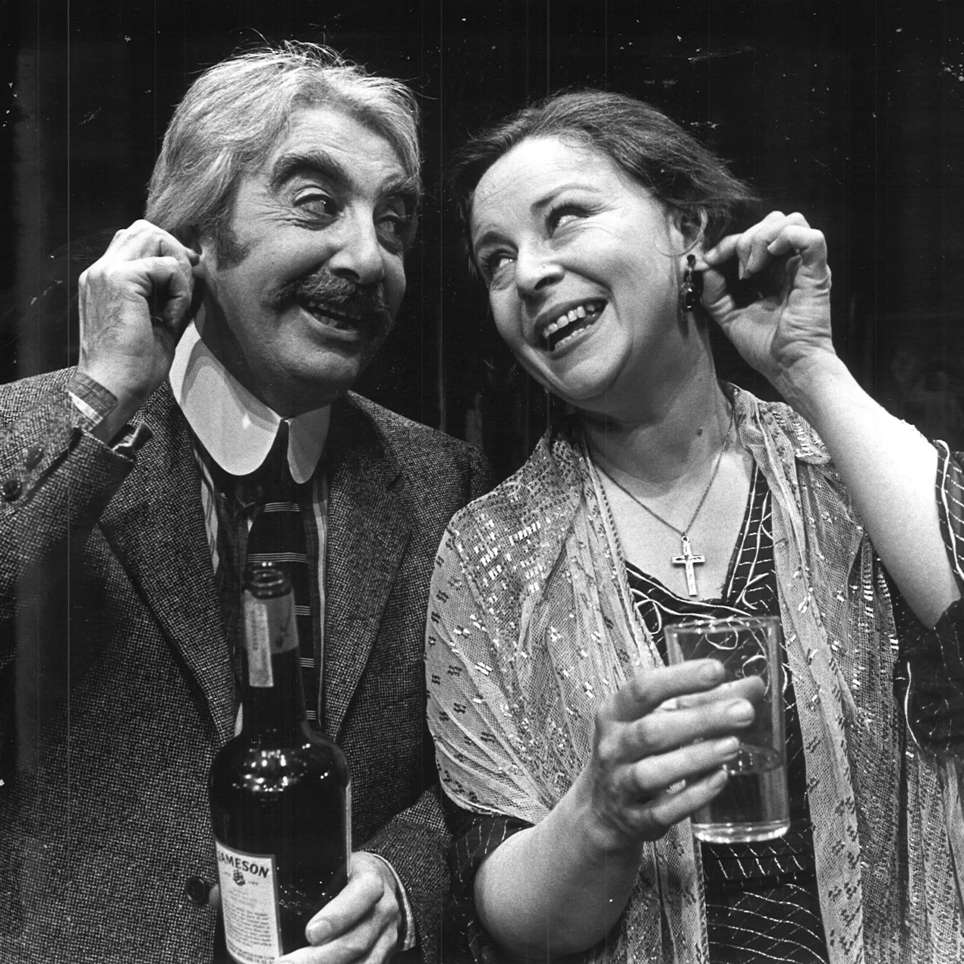 A man and a woman are seen standing together pinching they're ears and smiling while the man holds a bottle of wine and the woman holds a glass of water