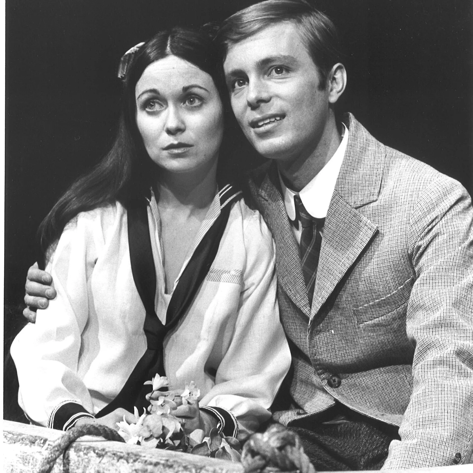 A woman dresses as a sailor and a man wearing a tuxedo are seen standing together and looking off into the distance