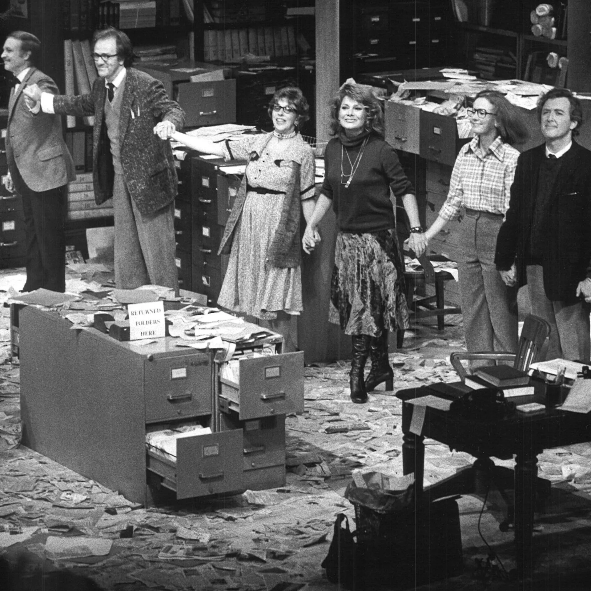 A group of people are seen standing together holding hands in front of a filing cabinet with papers all over the ground.