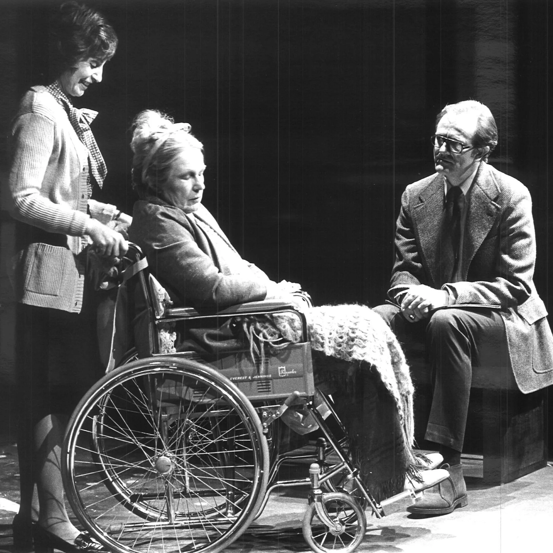 A woman is pushing another woman in a wheelchair whose eyes are closed while a man is sitting in front of them.