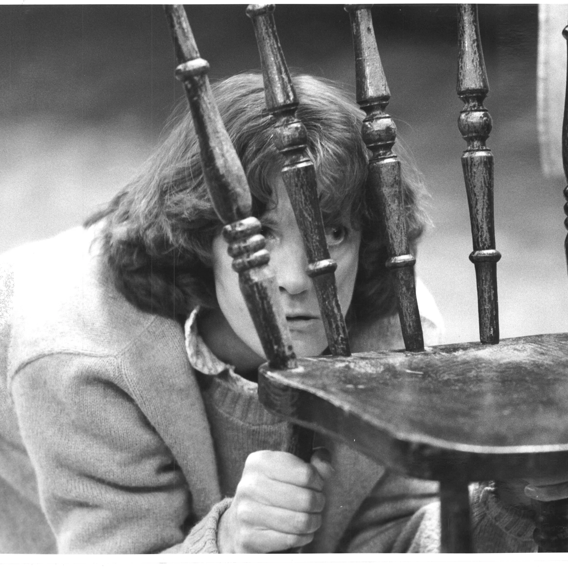 A woman is seen hiding behind a wooden chair and peeking out.