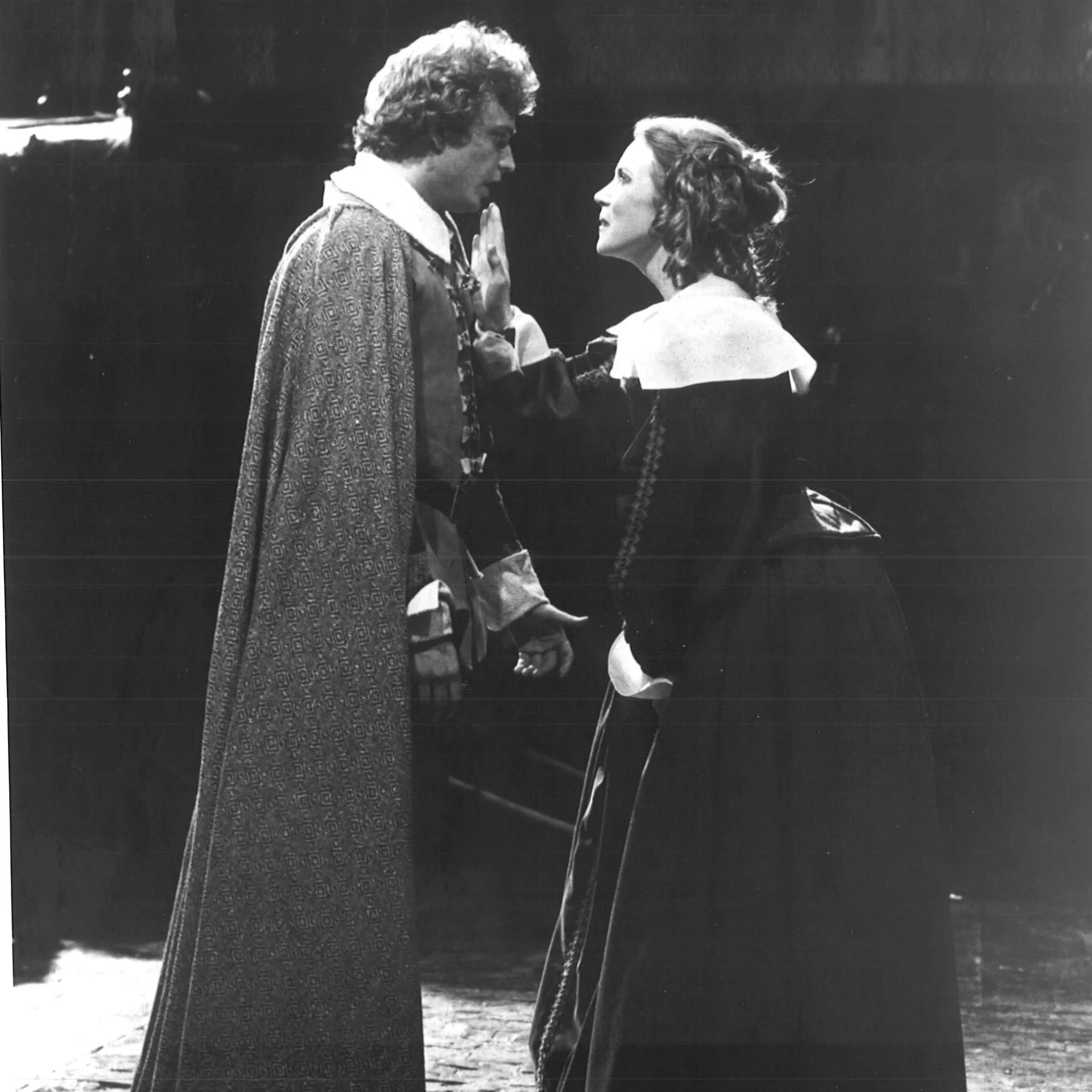 A man and woman are standing together talking and the woman has her hand on the man's chest.