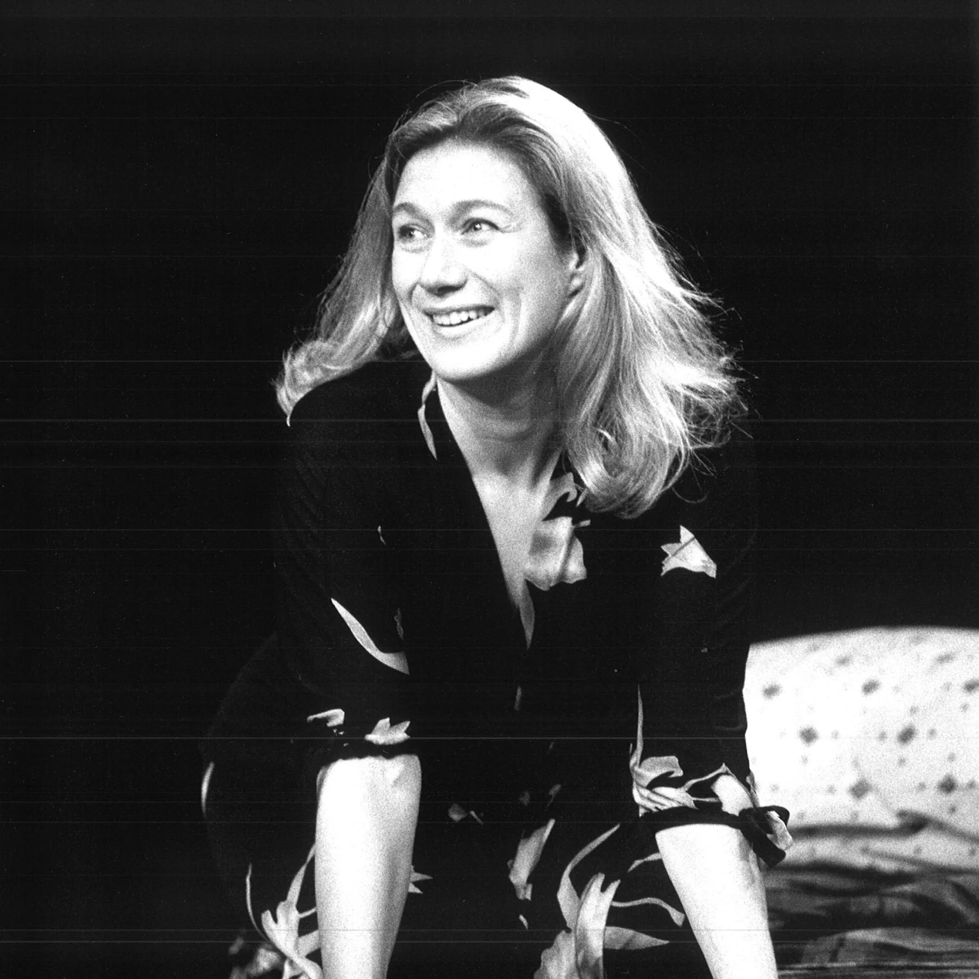 A woman perched on a bed looks off to the left smiling