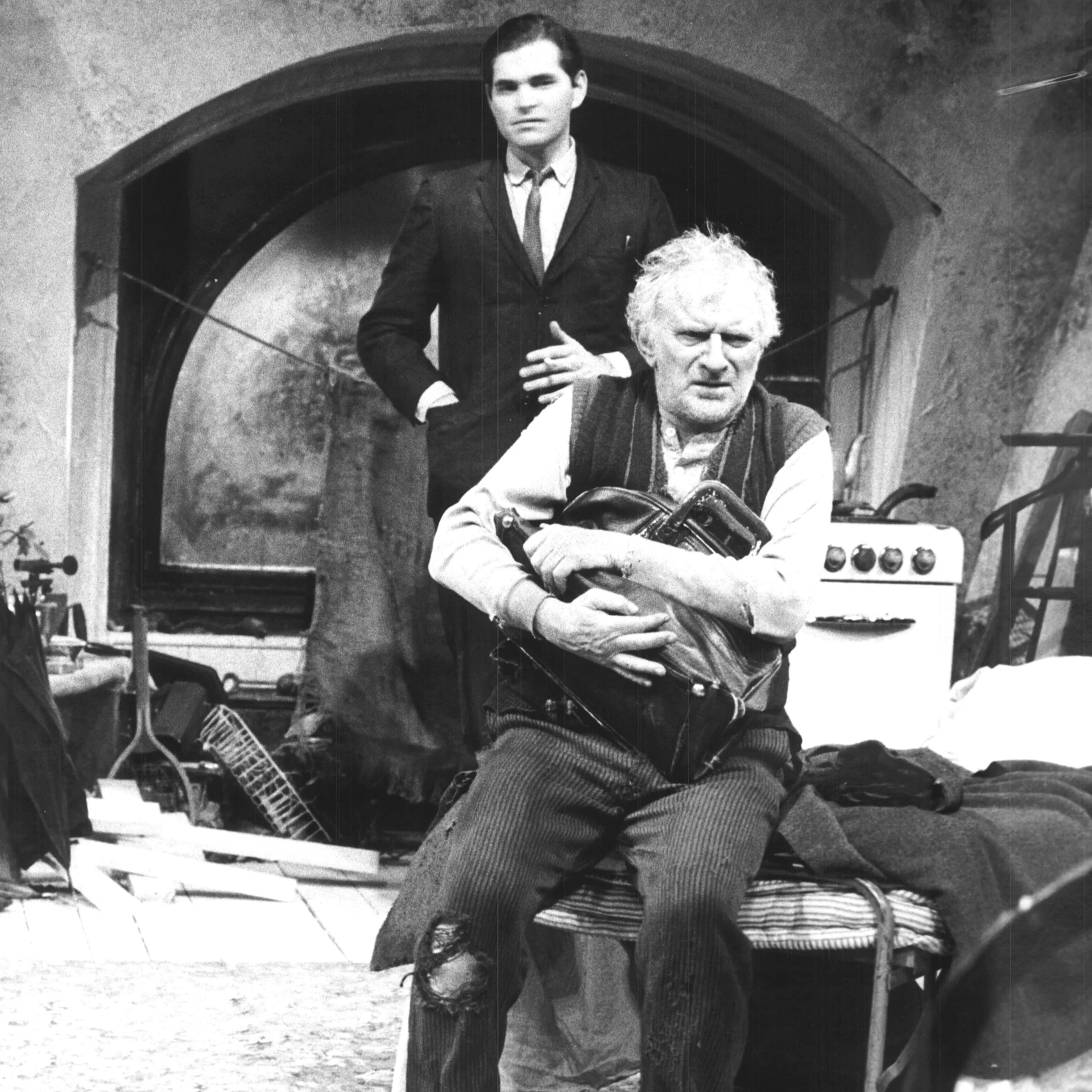 A man is seen sitting down and frowning while he clutches his bag and another man is behind him watching.