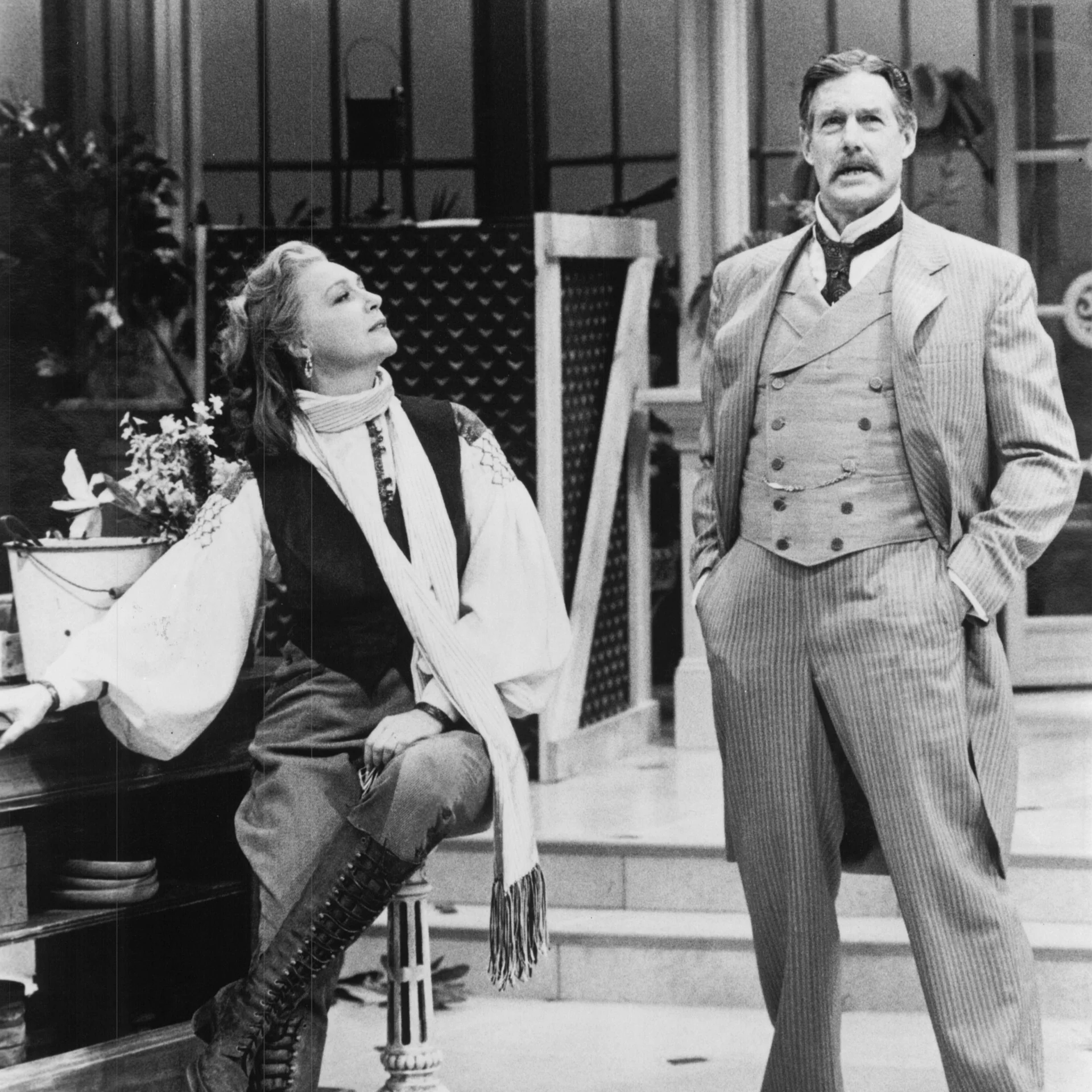 A woman sitting on a stool in high laced boots looks to her left at a man standing with hands in his pockets