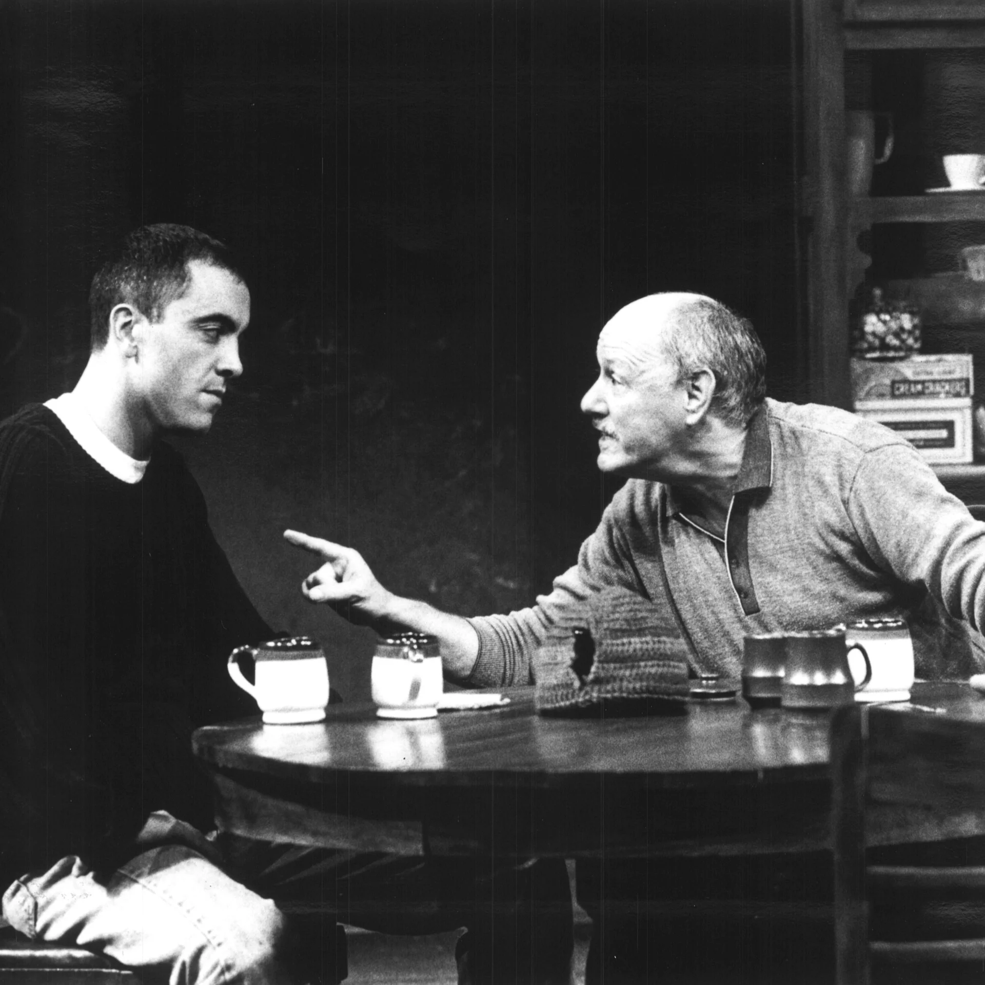 An older man sitting a table points his finger at the man sitting next to him