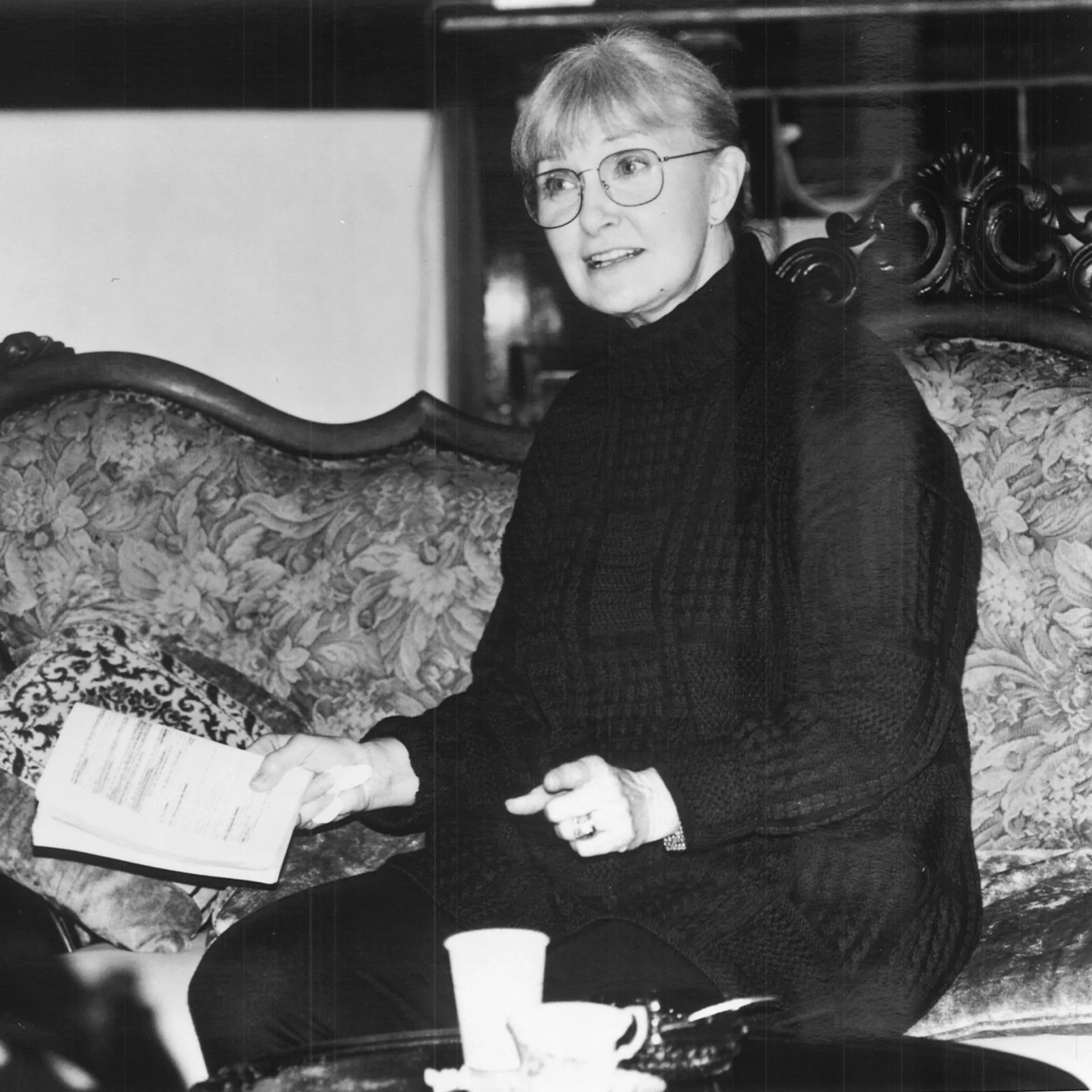 An older woman with glasses sits on a sofa holding a small book