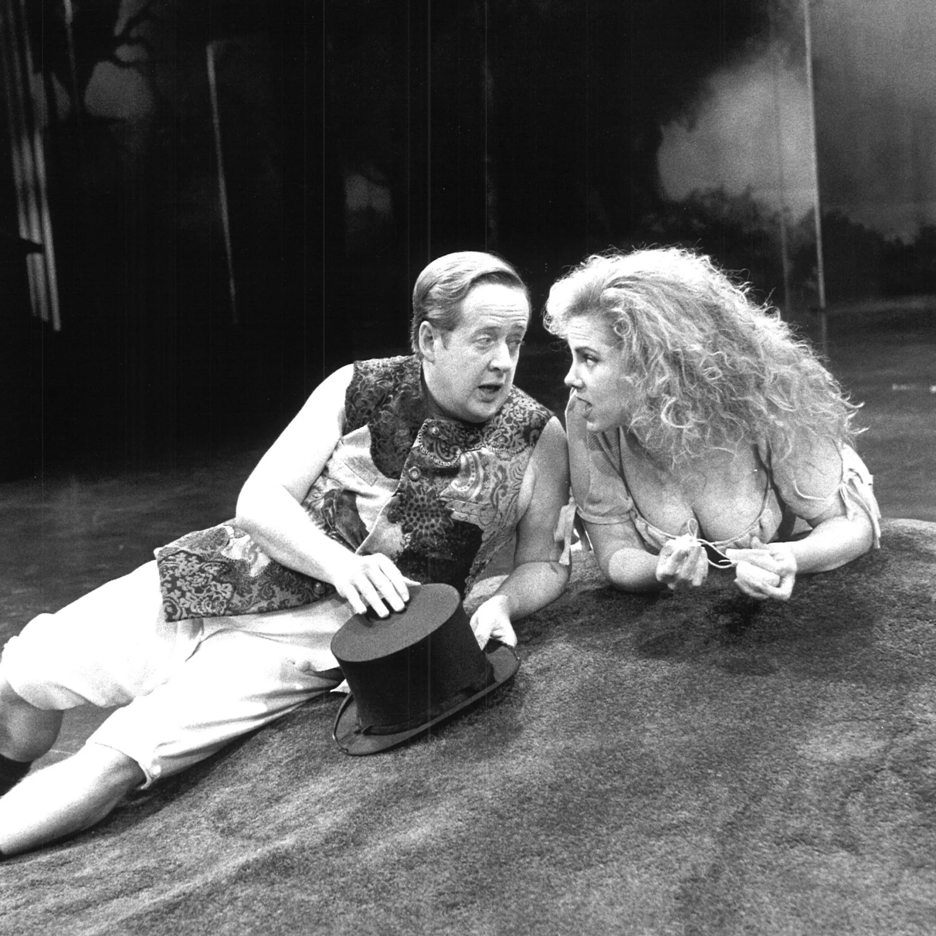 A man rests on his elbows holding a top hat, a woman lies on her front; they are chatting.