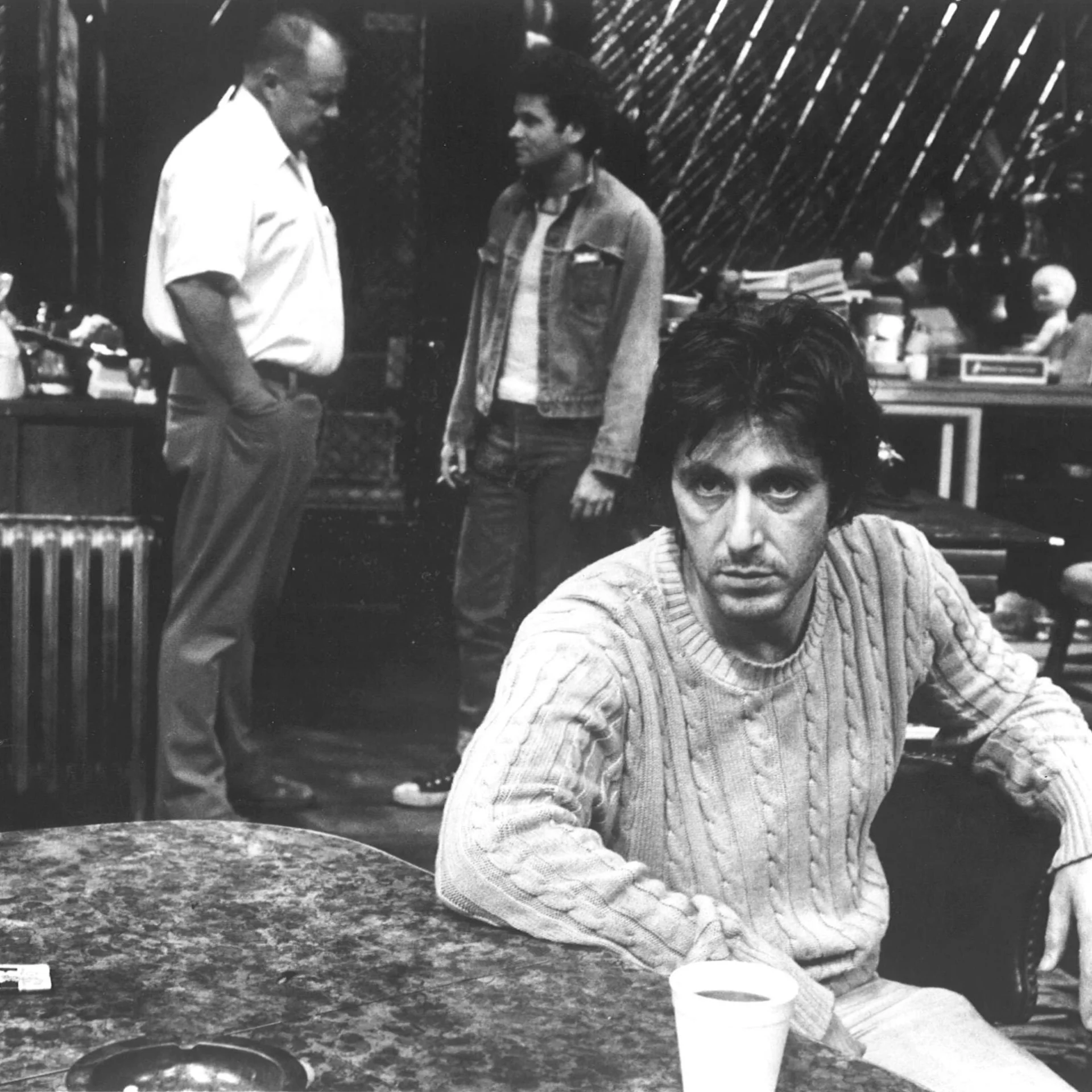 A man is sitting at a table looking off into the distance while two man are standing in the background talking