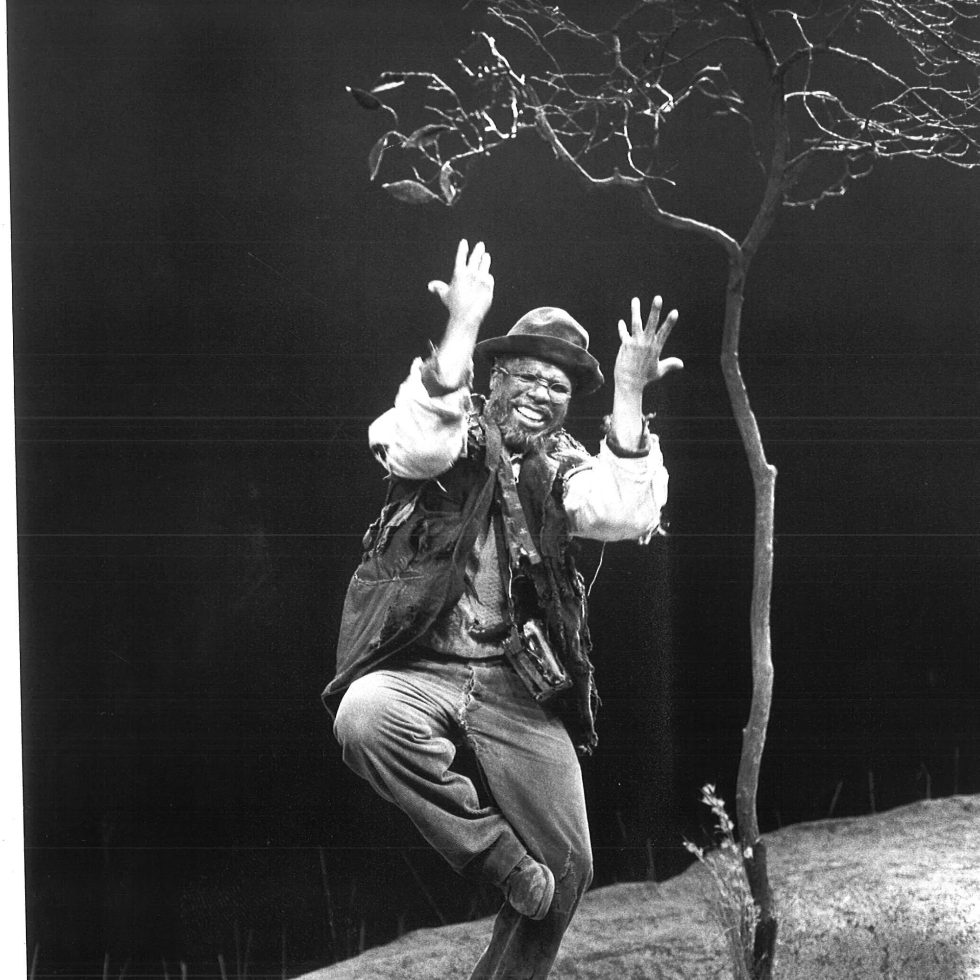 A man is seen standing on one leg and putting his hands up while he's smiling.