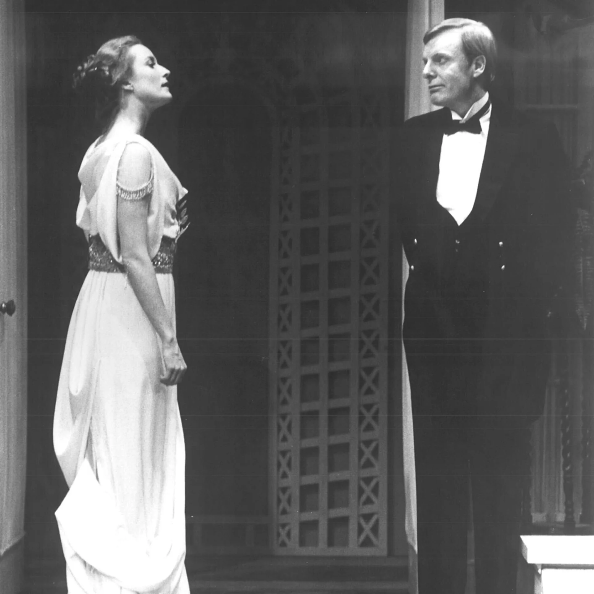 A woman wearing a white dress and a man wearing a tux are standing across from each other and talking