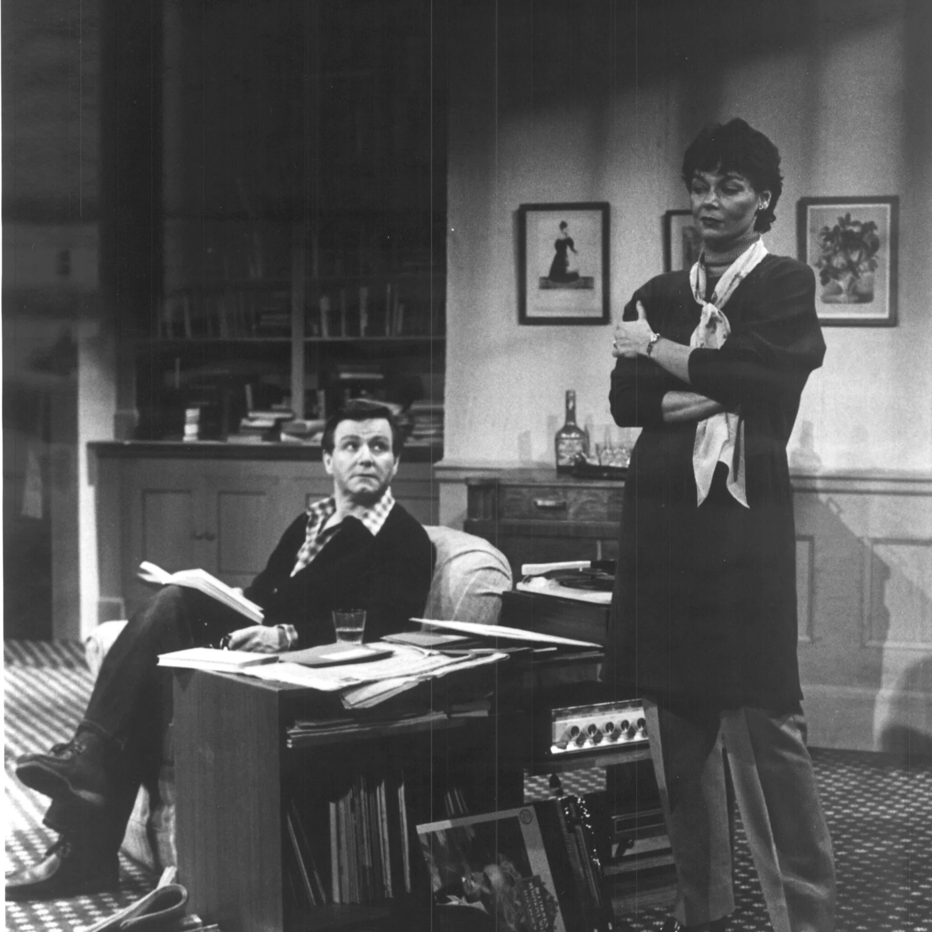 A man is seen sitting on the couch reading the paper while a woman is standing next to him crossing her arms.