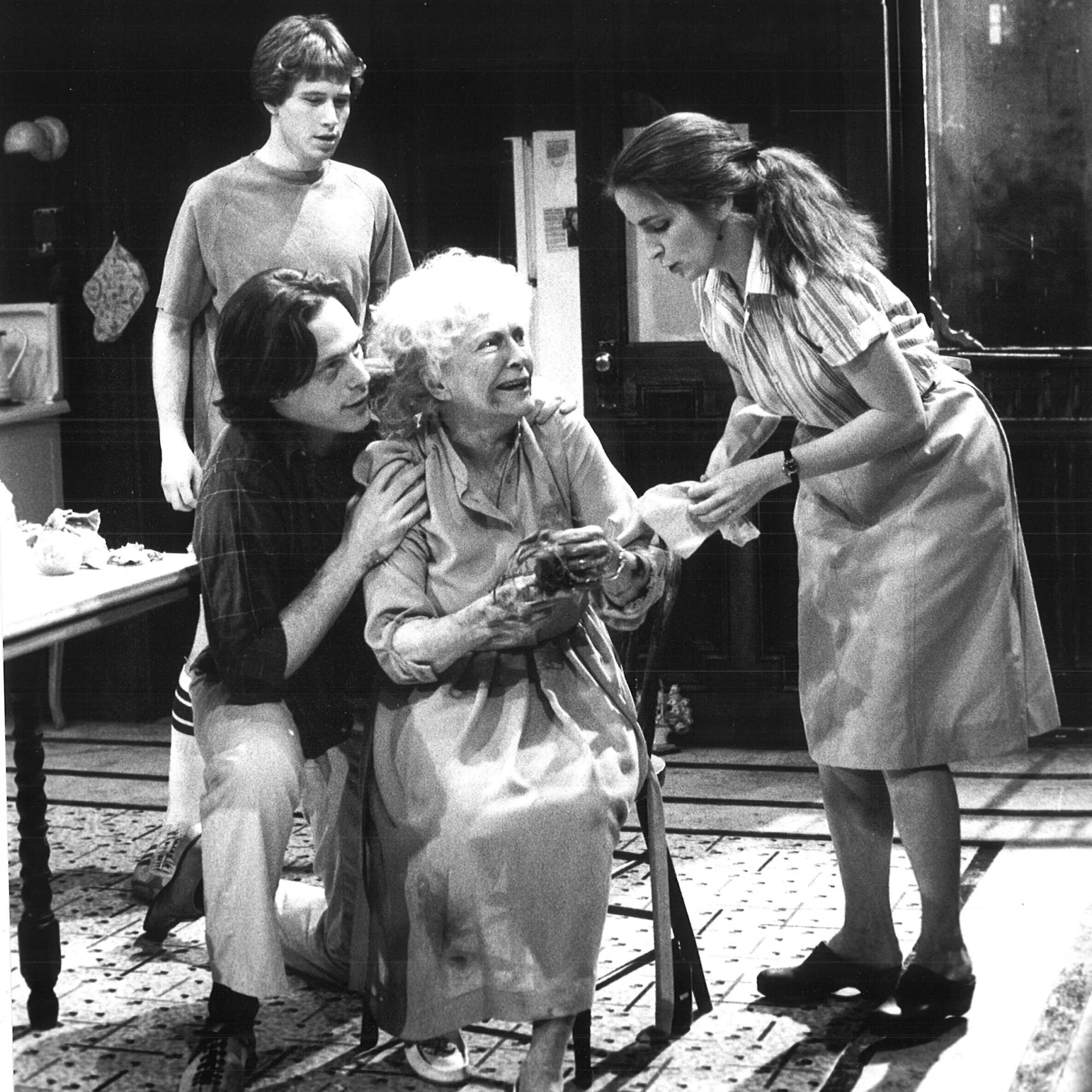 An old woman is seen sitting in a chair and another woman is handing her something while two men stand behind them.