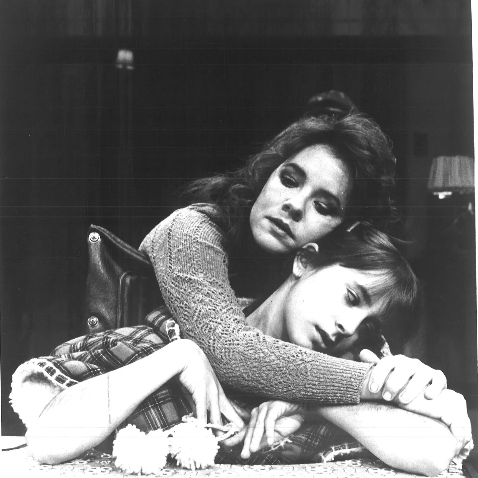 A girl is sitting at a table while a woman is hugging her from behind.