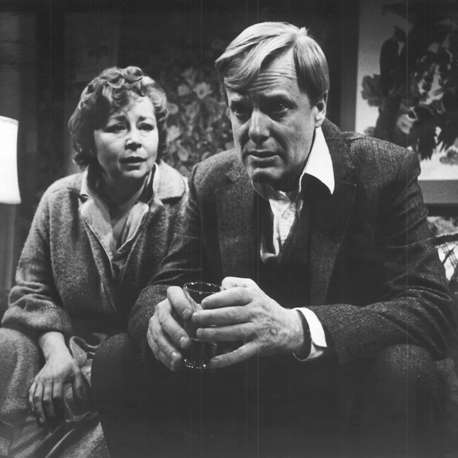 A man and woman are sitting on a couch and the man is leaning forward holding a cup.