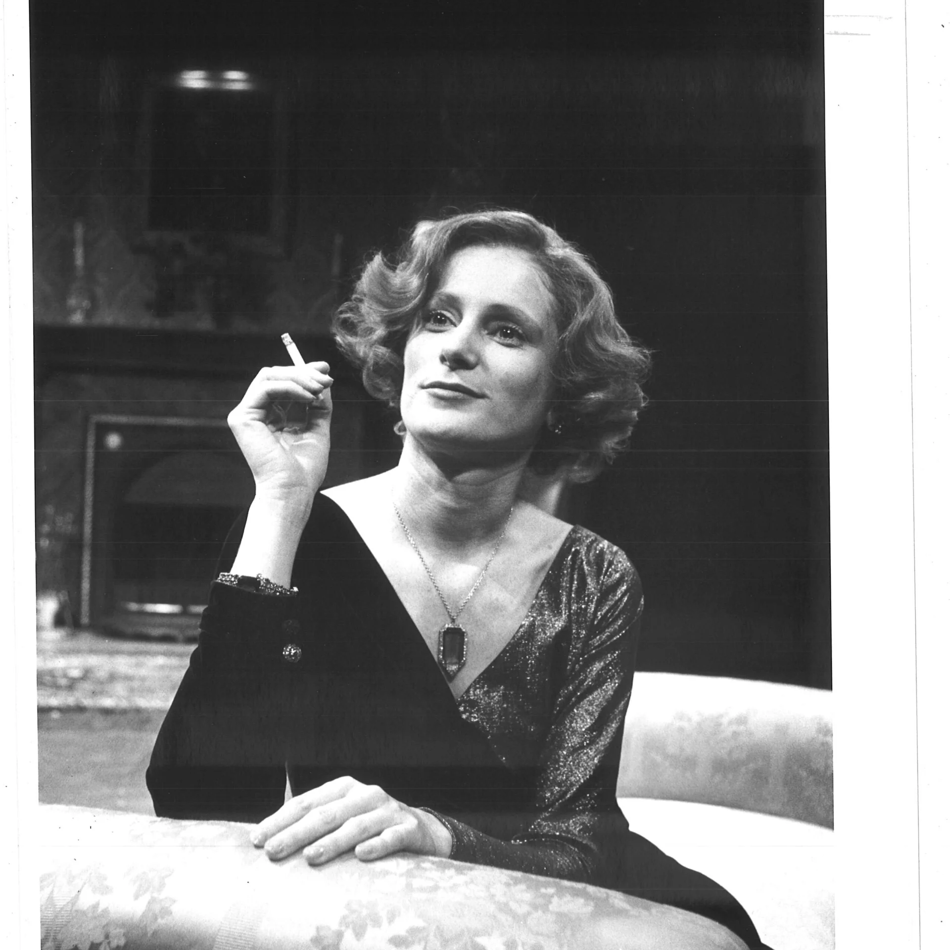 A woman in sitting on a coach smirking while she smokes a cigarette.