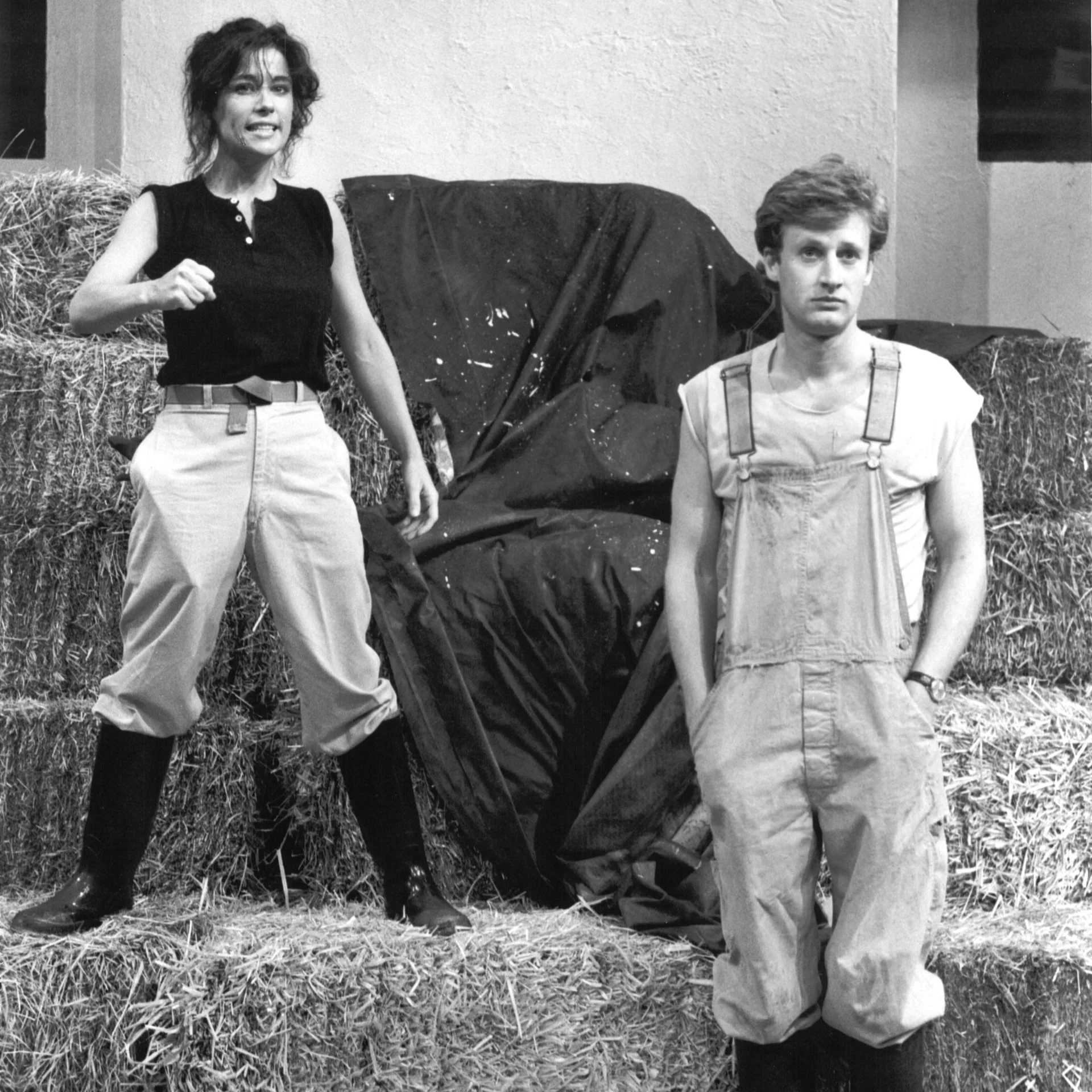 A woman is standing on a hay stack while a man stands next to her in overalls.