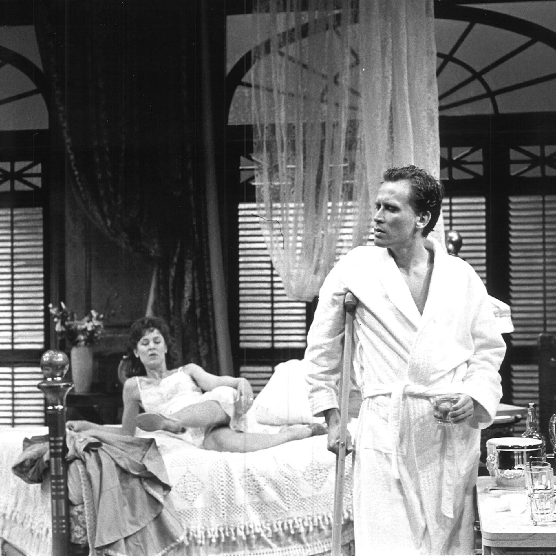 A woman is laying on a bed in lingerie while a man is standing on the edge of it wearing a bathrobe.