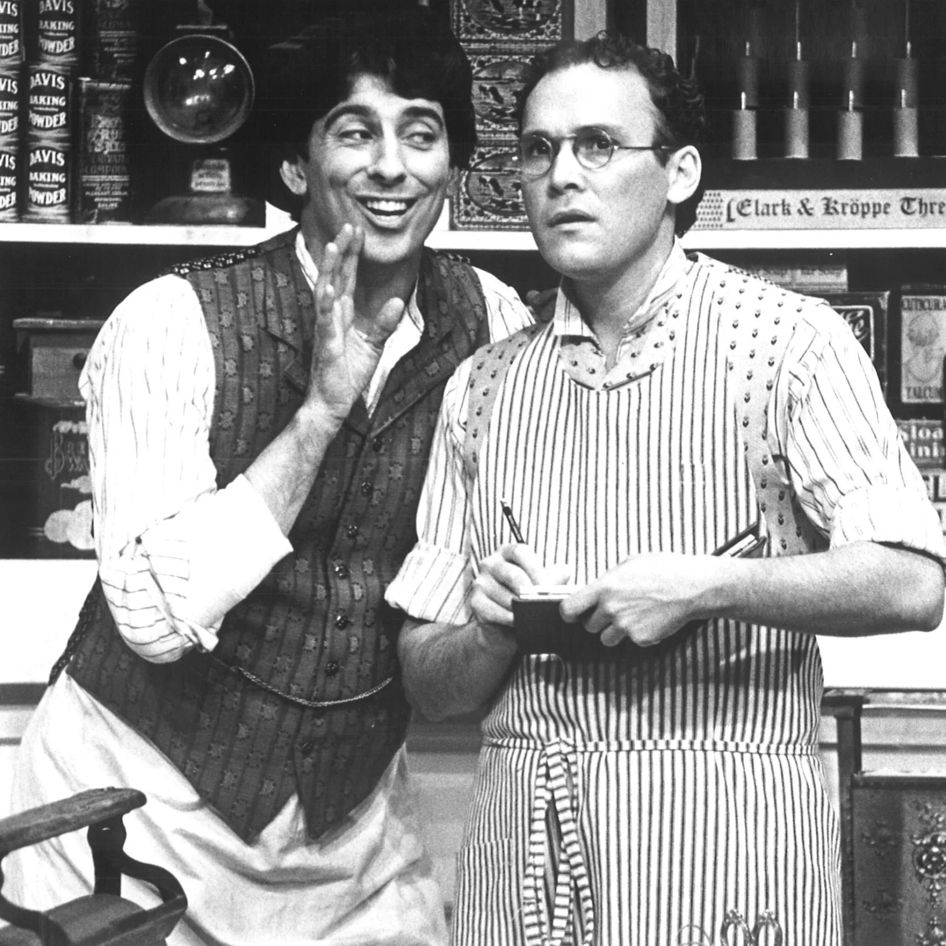 Two men are standing behind a store counter and one of them is holding his hand up and talking while the other is listening.