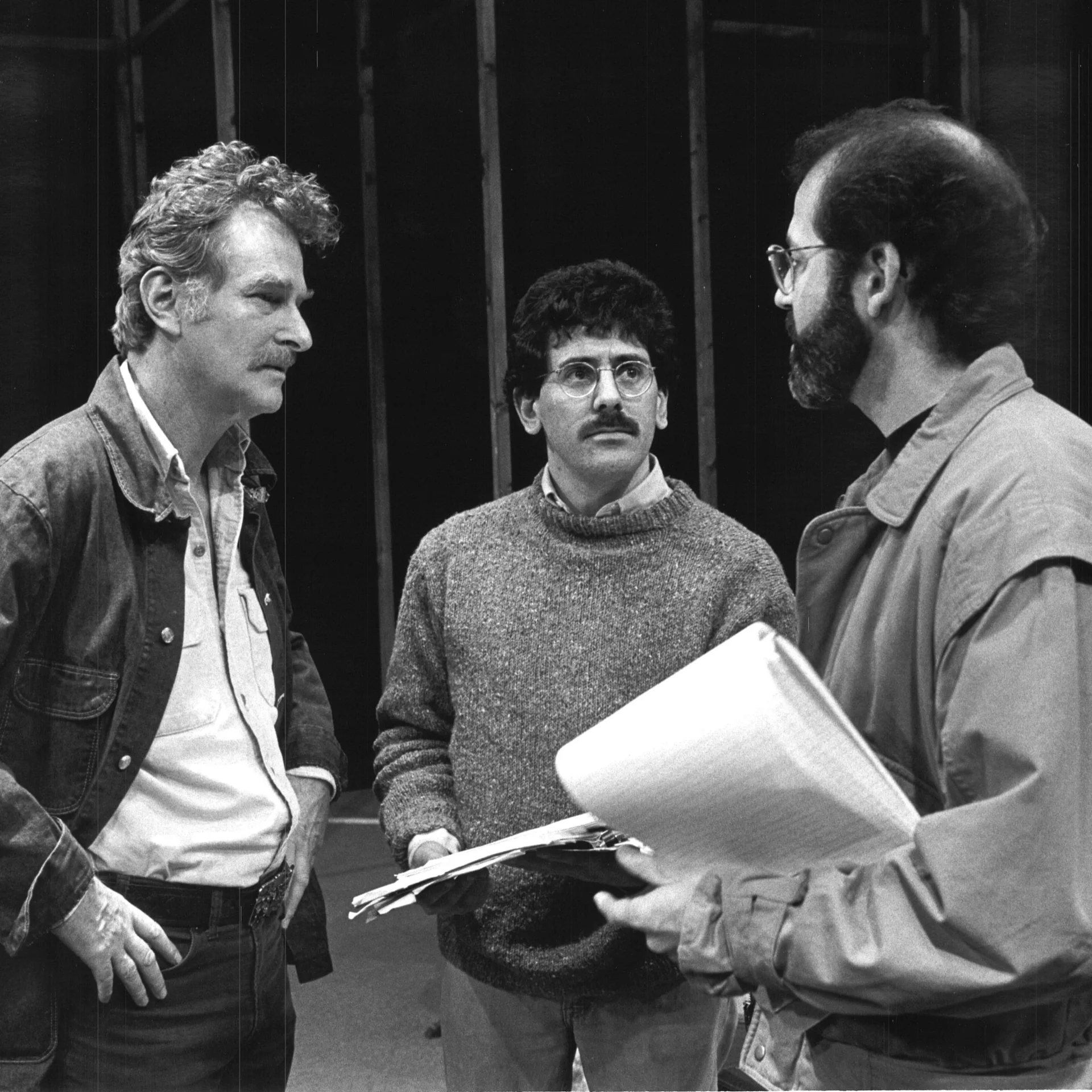 Three men are standing together holding notebooks and having a conversation.