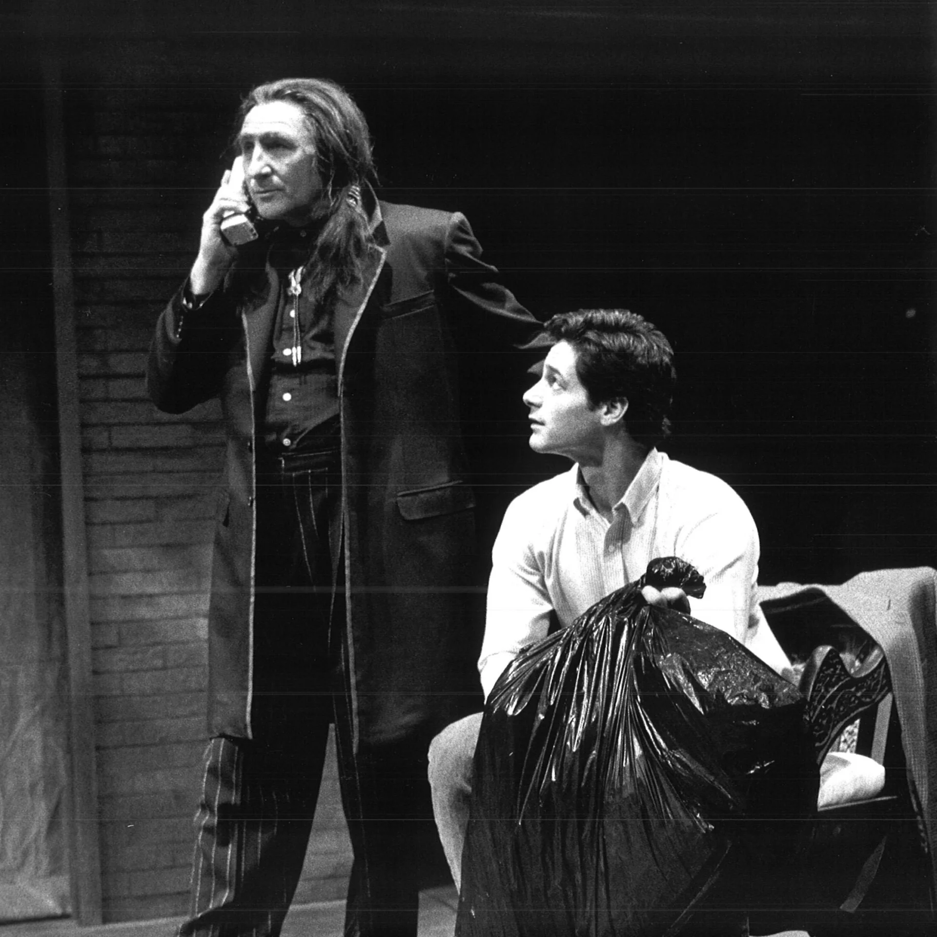 A man sits with a trash bag looking up at a man holding a phone