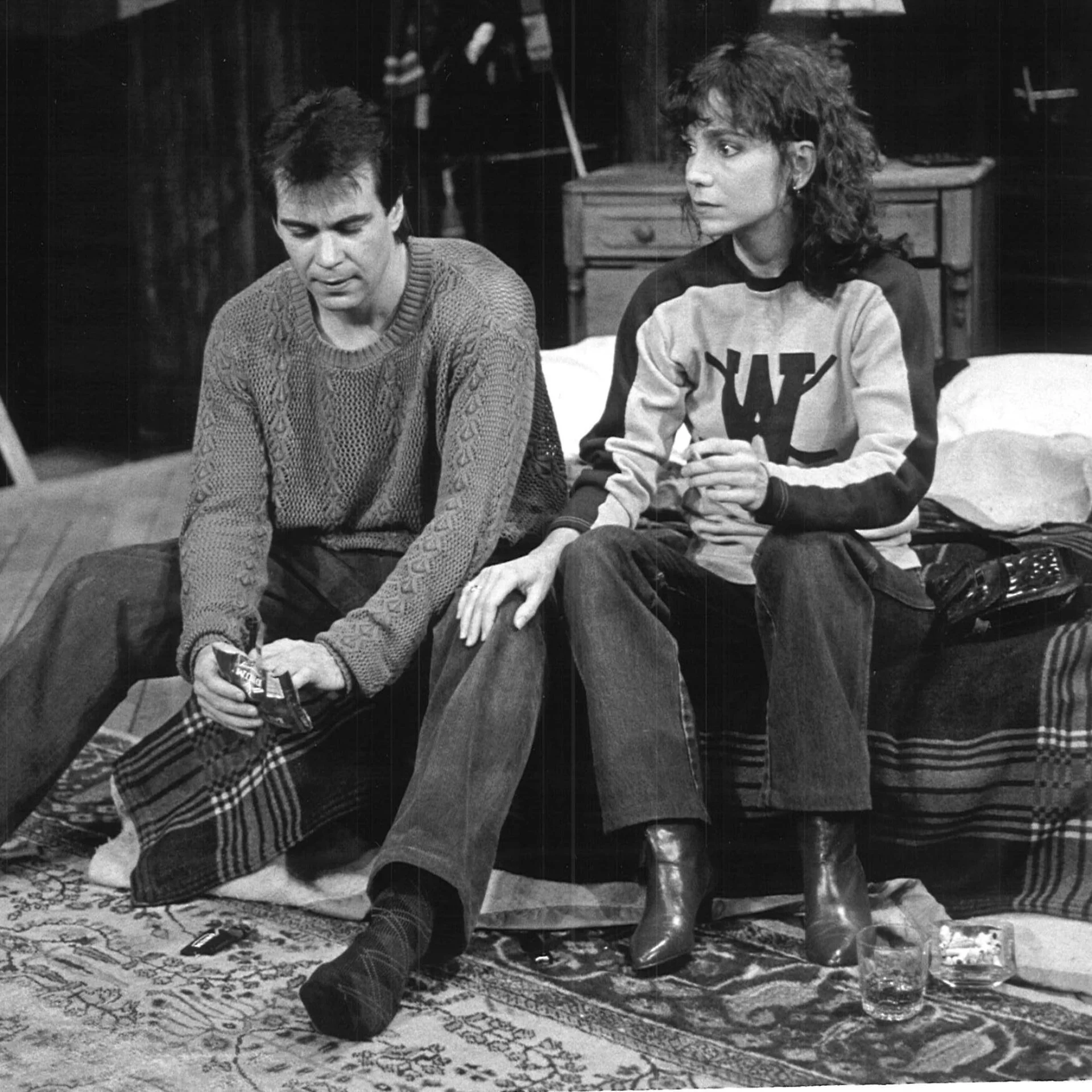 A woman and man are sitting together on the edge of a bed and the woman has her hand on his leg.