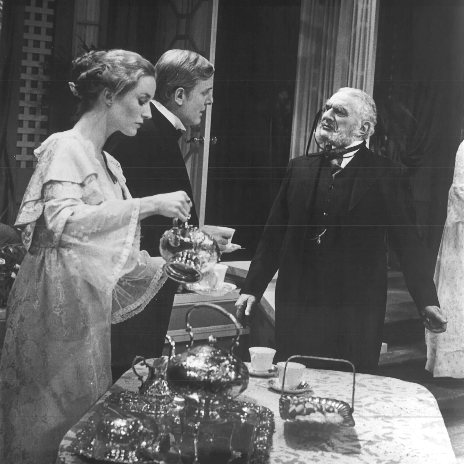 A woman is pouring herself tea while two men are standing next to her talking.