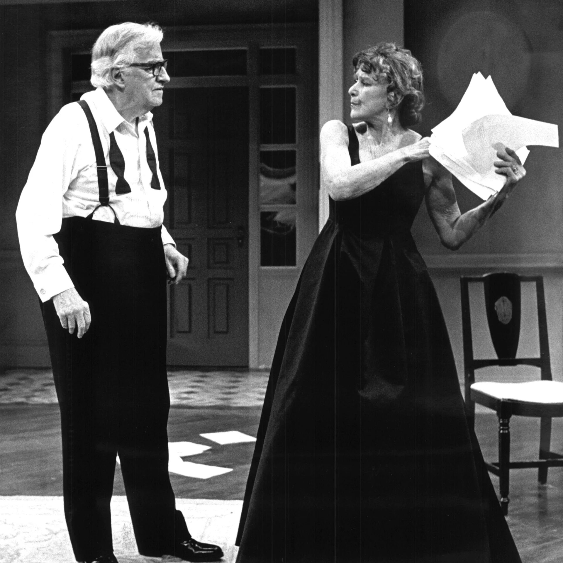 A Woman wearing a black dress is ripping papers away from a man wearing a bow tie and suspenders.