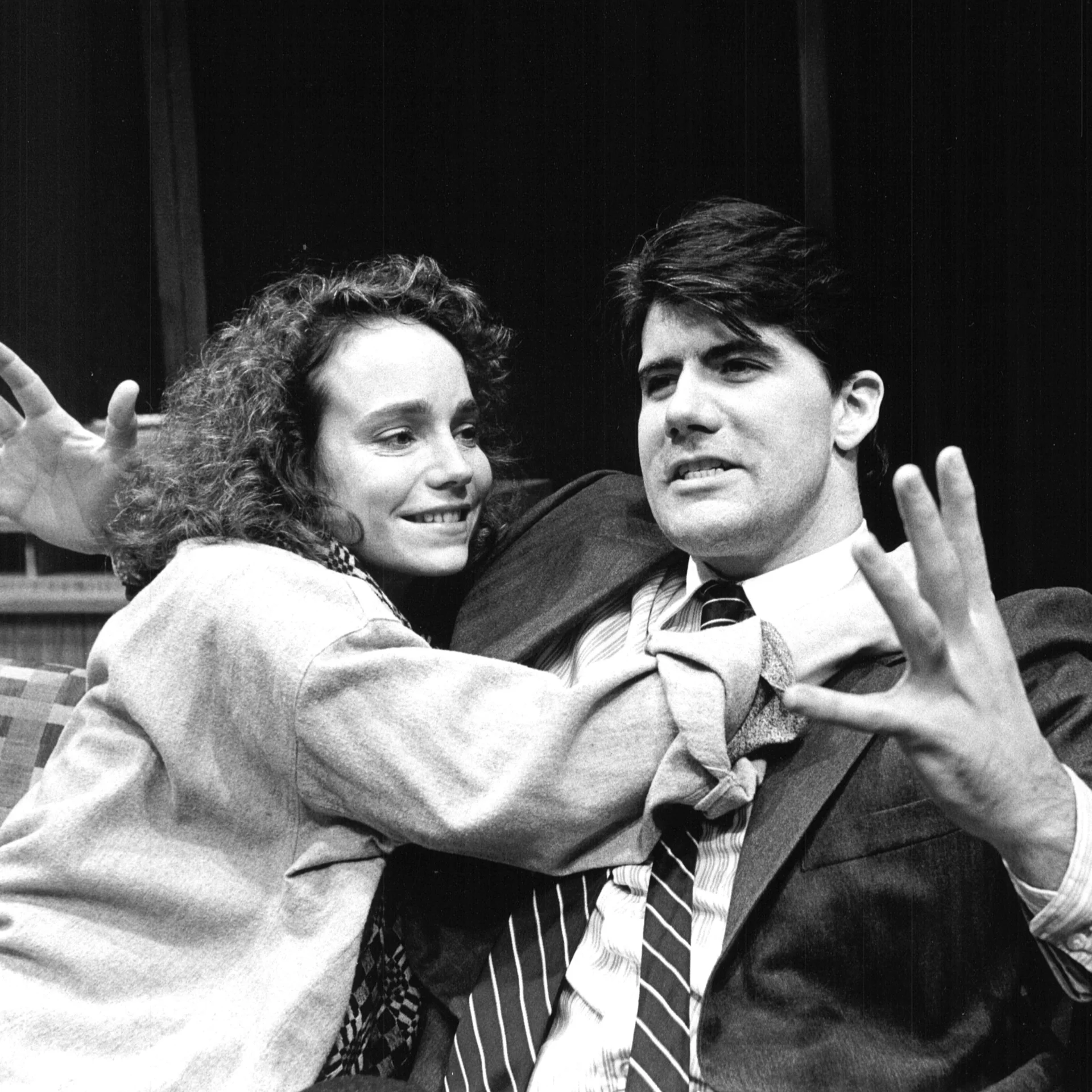 A man and a woman are sitting on a couch and the woman is trying to hug the man and he is throwing his hands up.