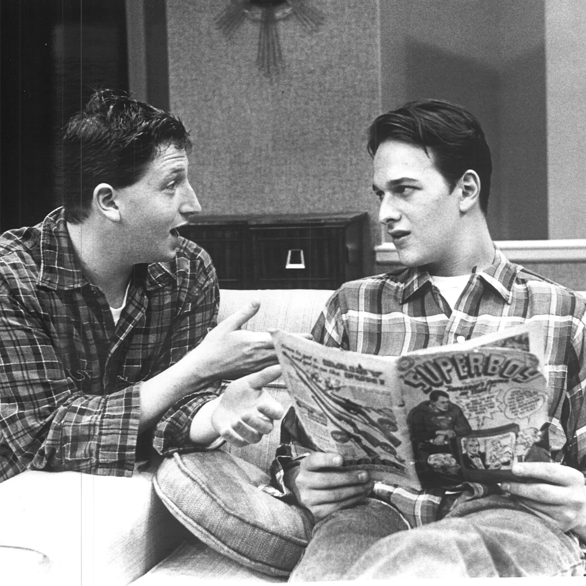 A man is sitting on a couch reading a newspaper while another man is trying to engage conversation with him.