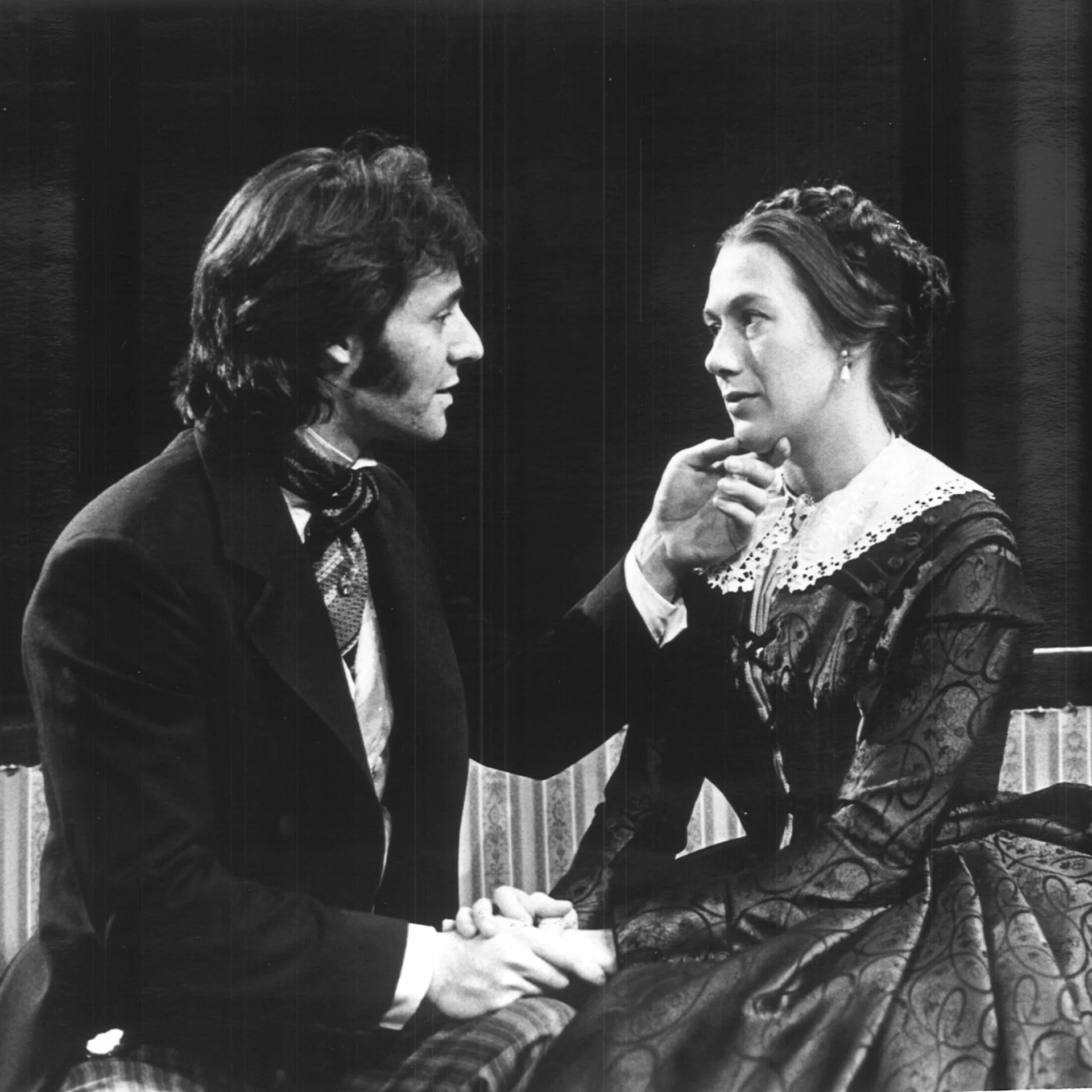 A woman wearing a Victorian dress is sitting on a couch with a man in a suit who is holding her chin.