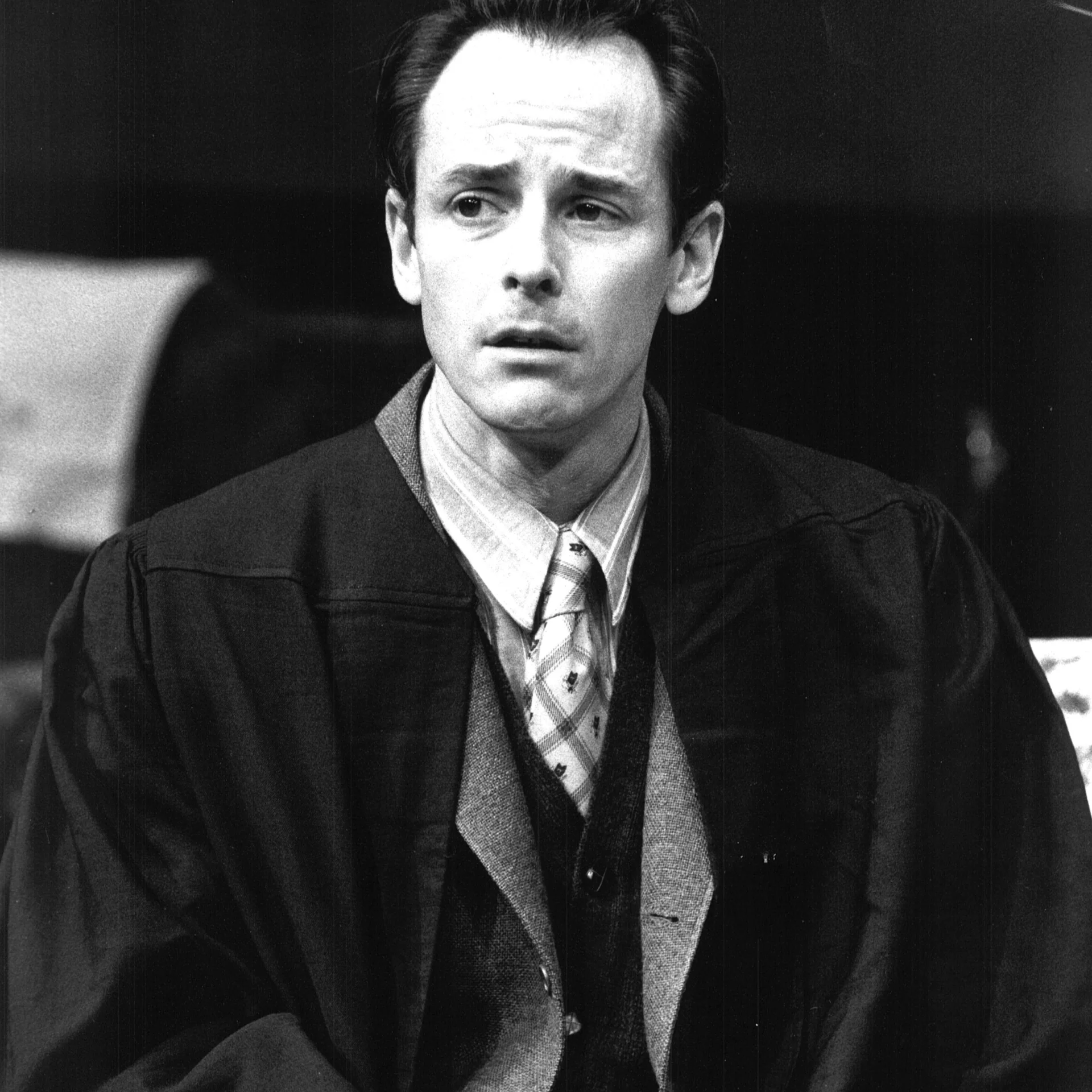 A man is sitting wearing a trench coat and staring off into the distance.