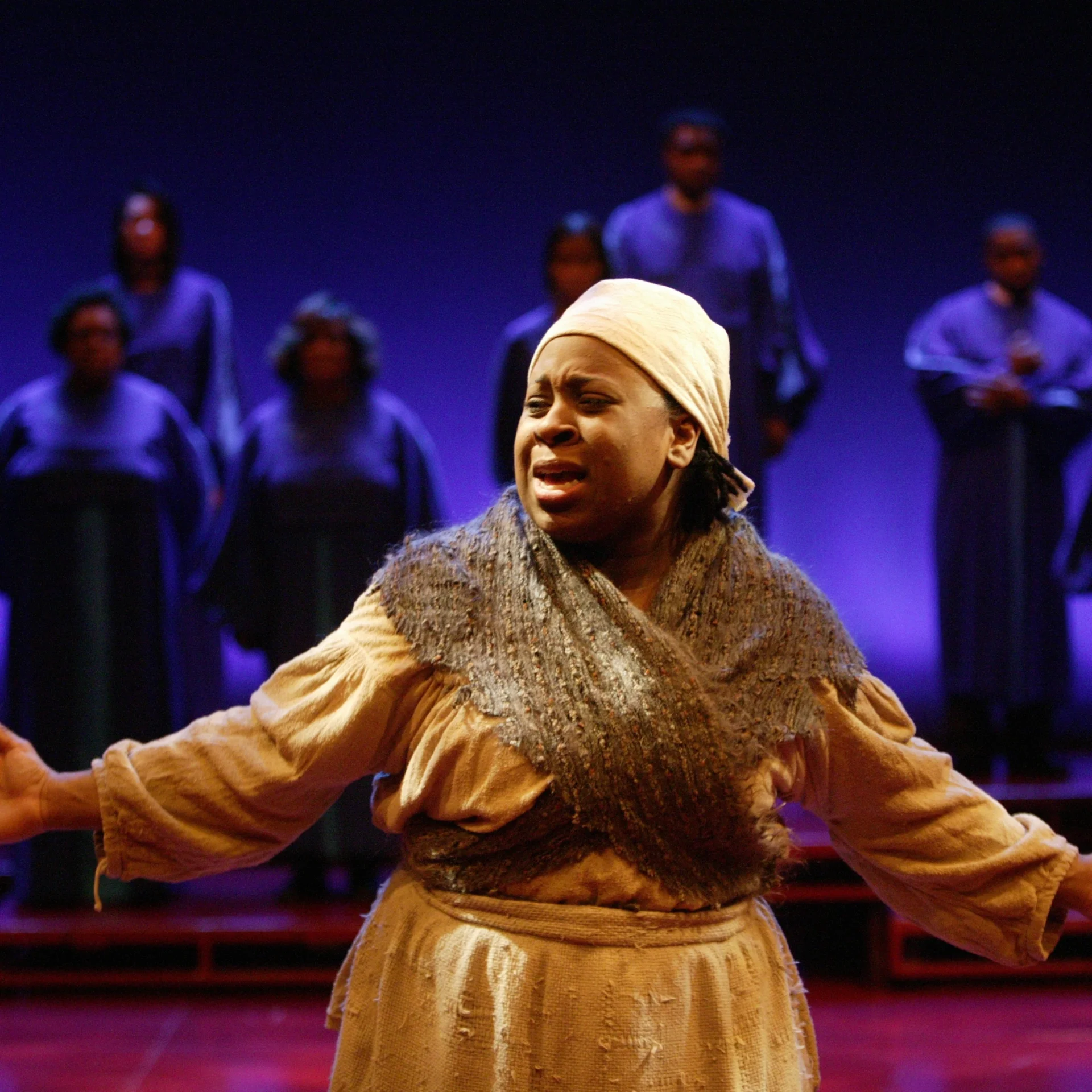 A Black woman stands with her arms spread with a scattering of choir singers in the background