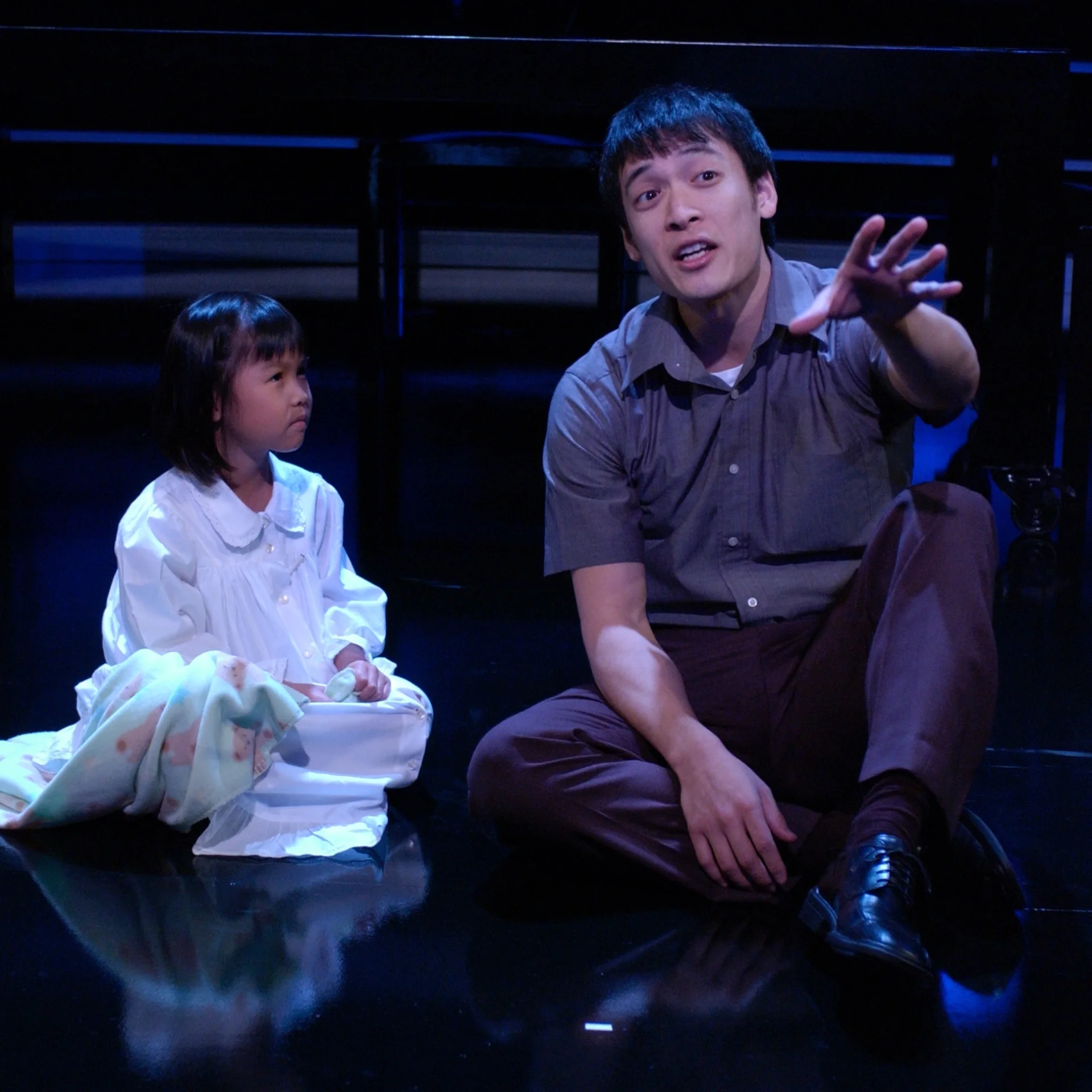 A man and little girl sit on the ground as the man talks out towards the audience