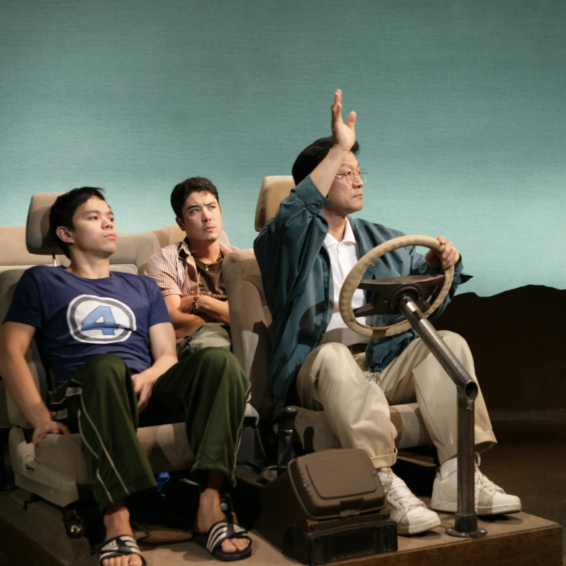 Three men sit in a makeshift car; the man driving is raising his arm