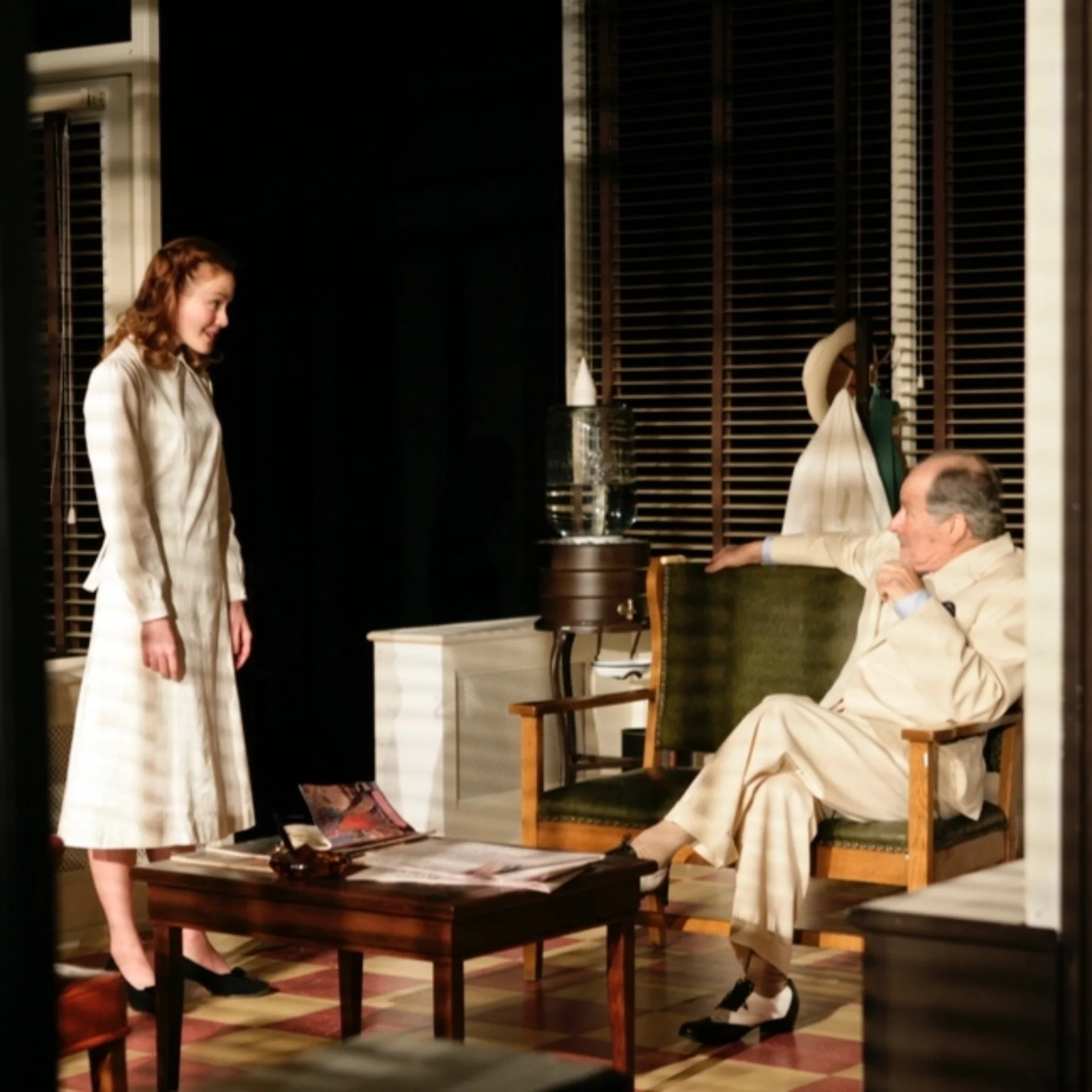 A young woman standing speaks to an older man sitting cross-legged in a living room
