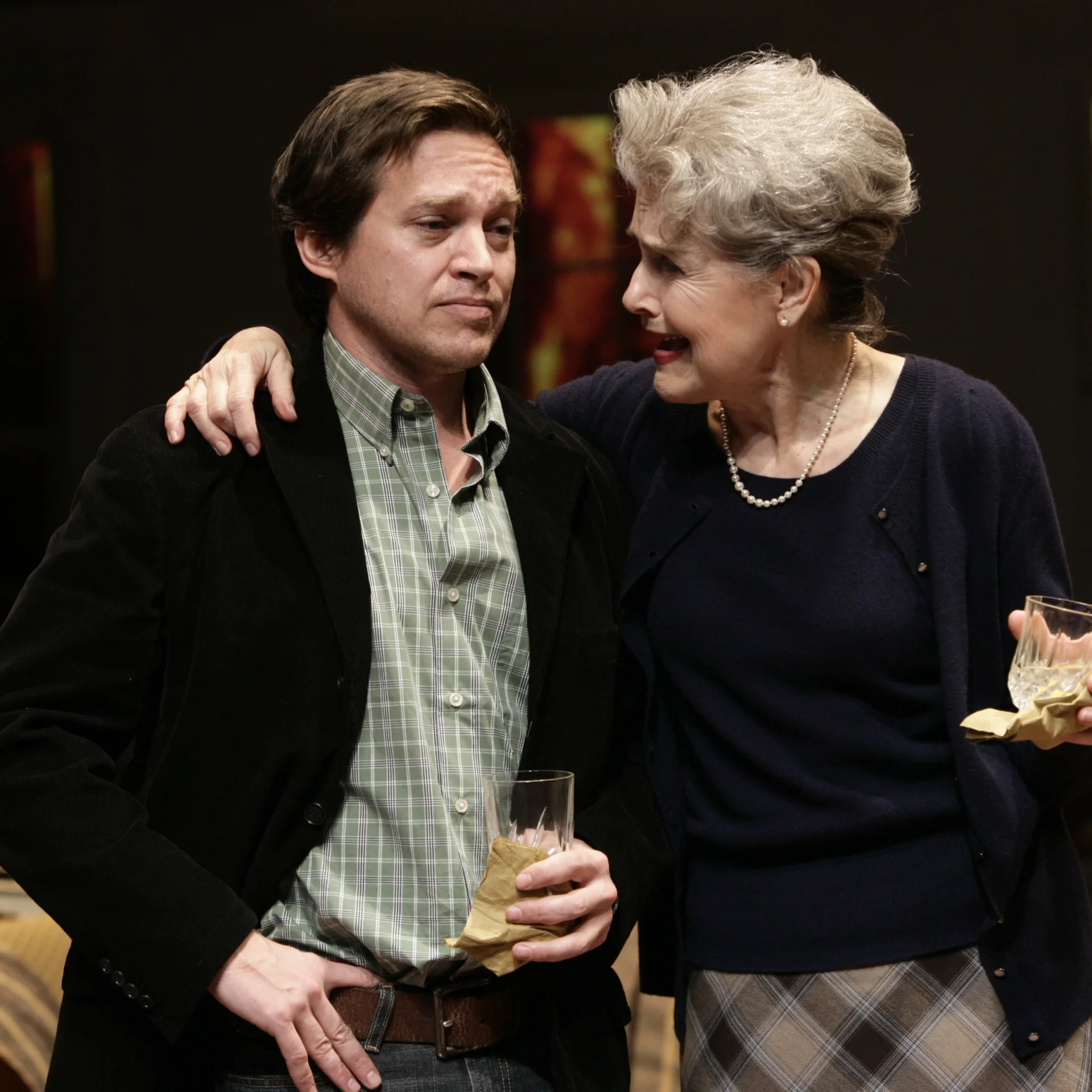 An older woman holding a drink is talking to another man holding a drink with her arm around his shoulders
