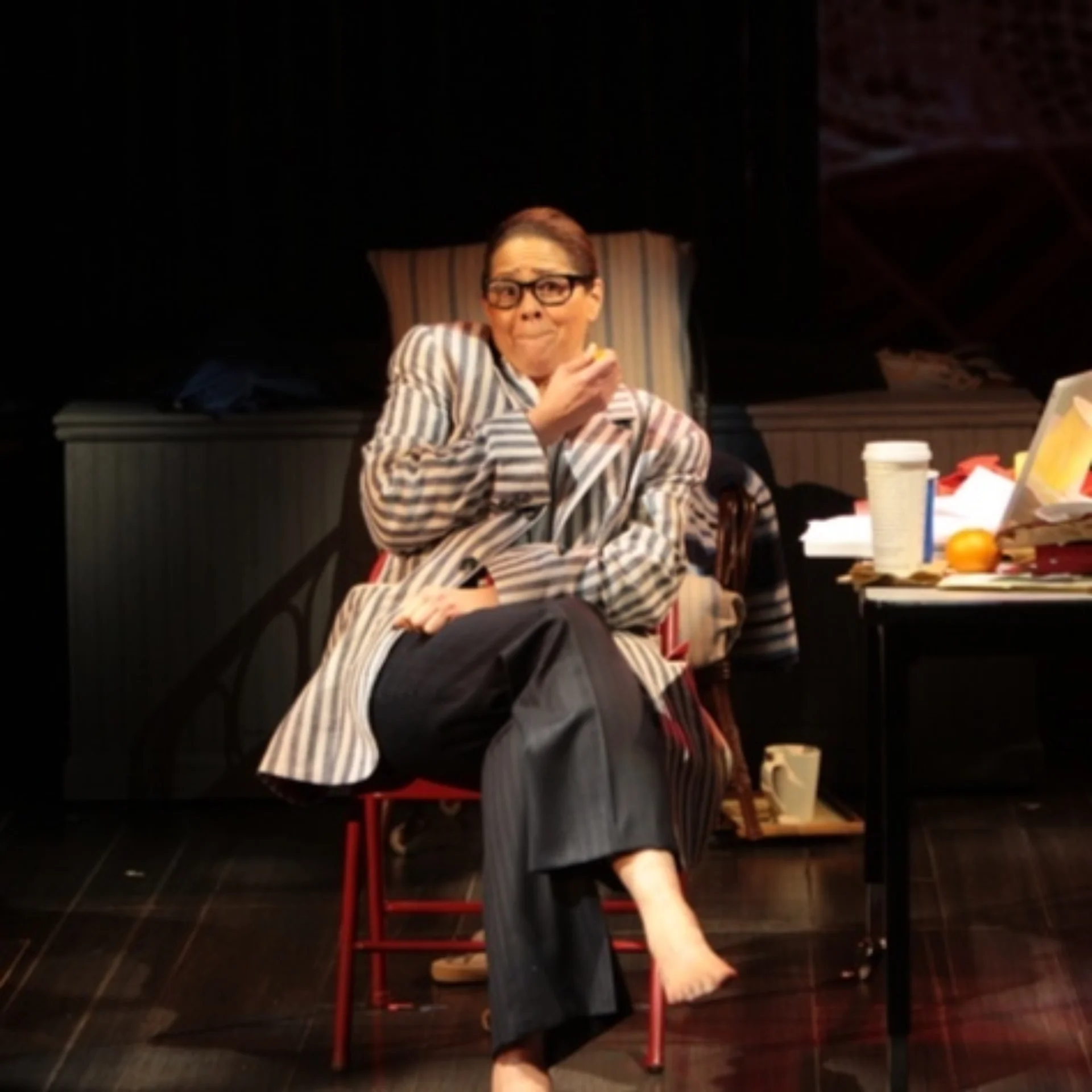 A woman in glasses sits cross-legged next to a messy table