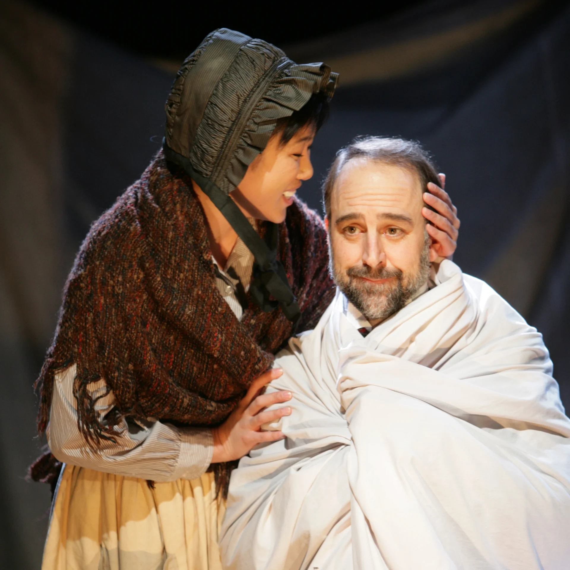 A woman in a bonnet holds a man wrapped in a sheet